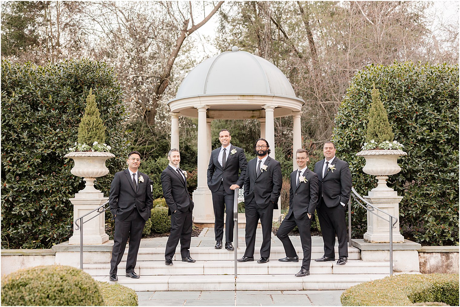 groom stands on steps in Park Chateau Estate gardens with groomsmen in black suits