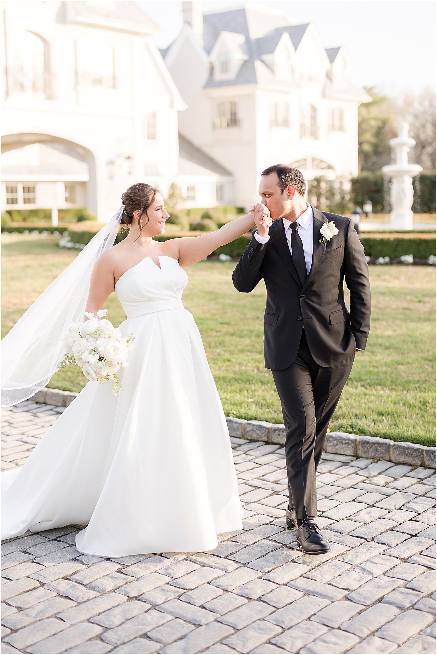 groom in black suit kisses bride's hand walking near Park Chateau Estate