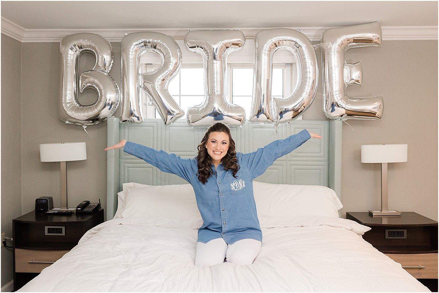 bride kneels on bed in button up shirt under BRIDE balloons 