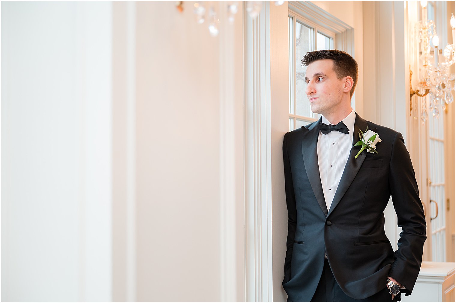 groom leans against white wall by window at Park Savoy Estate