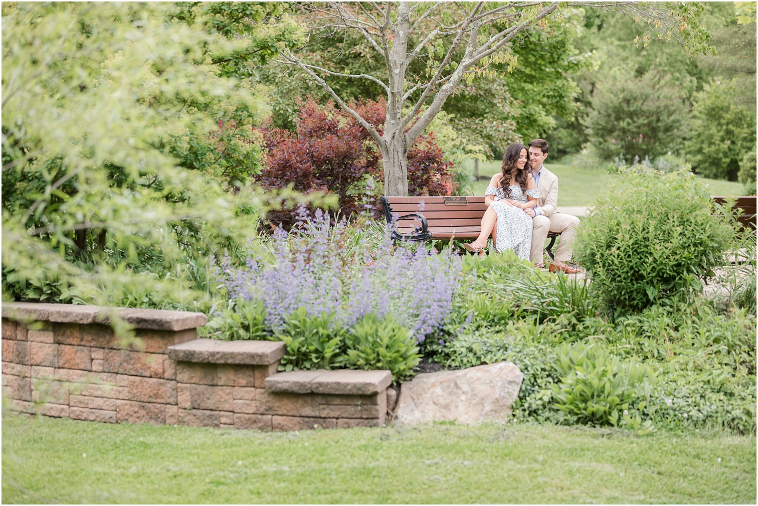 engaged couple sits on wooden bench behind purple and green flowers