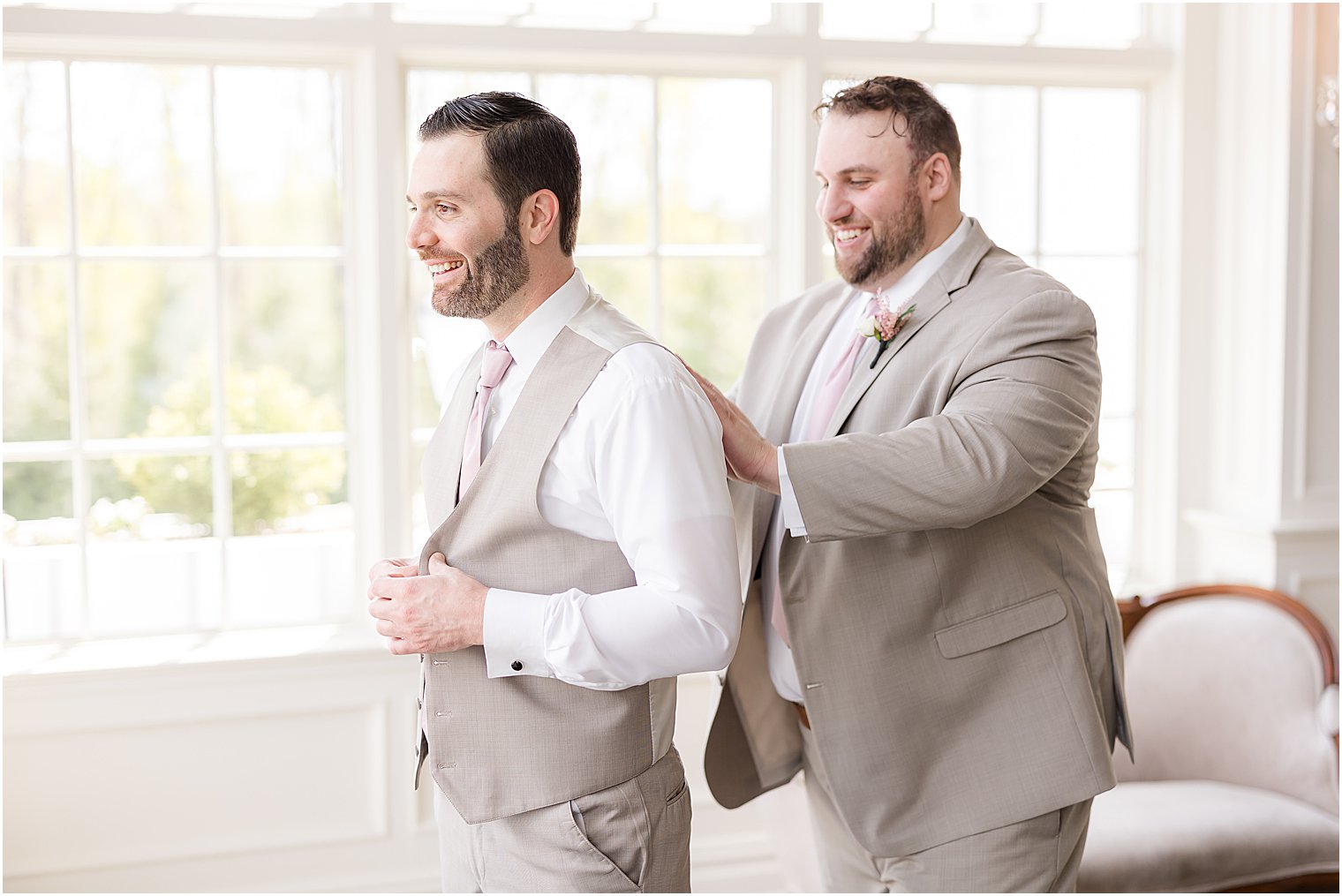 groomsman in tan suit helps groom into vest for spring wedding
