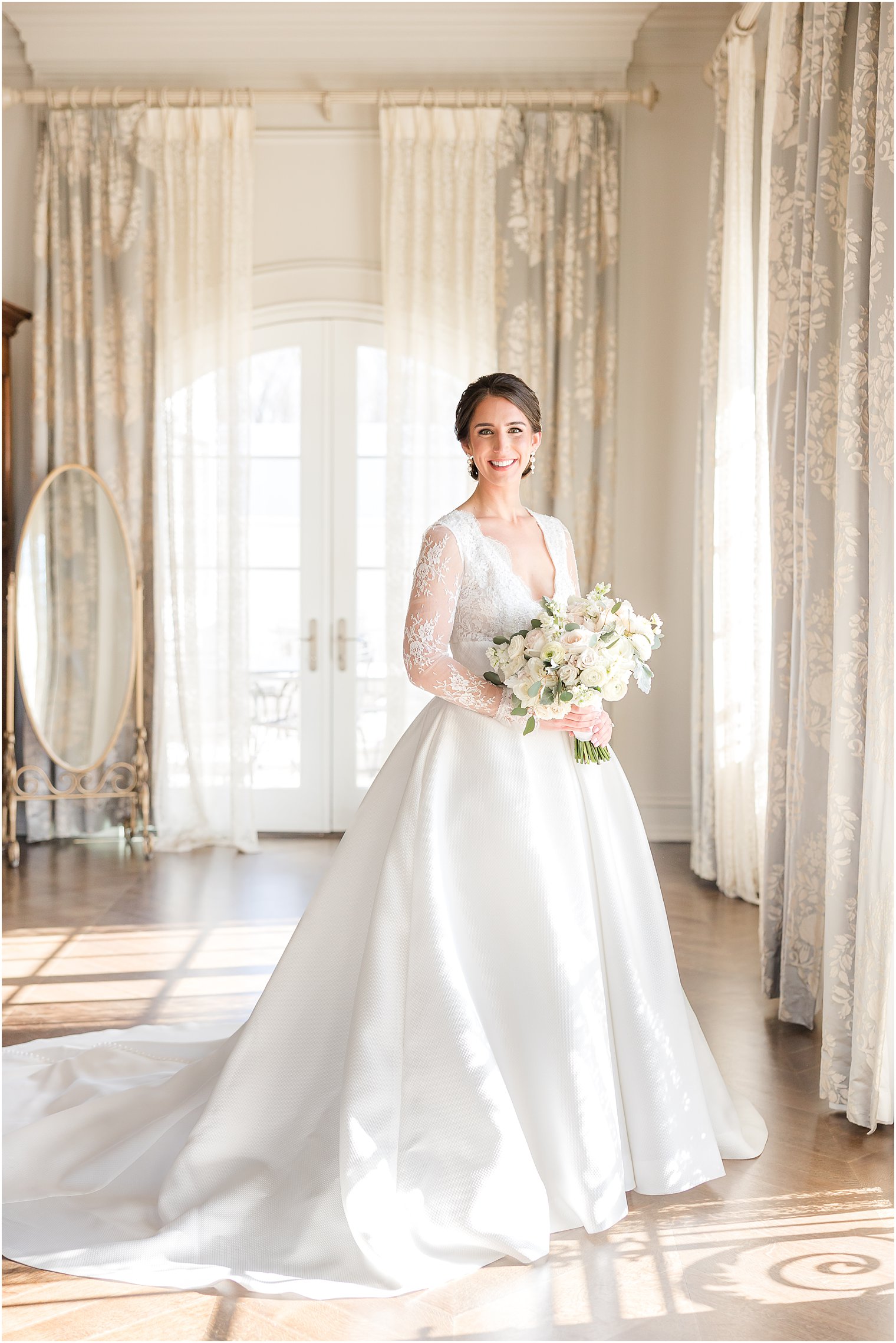 bride stands in wedding dress with lace sleeves by open window at Park Chateau Estate 