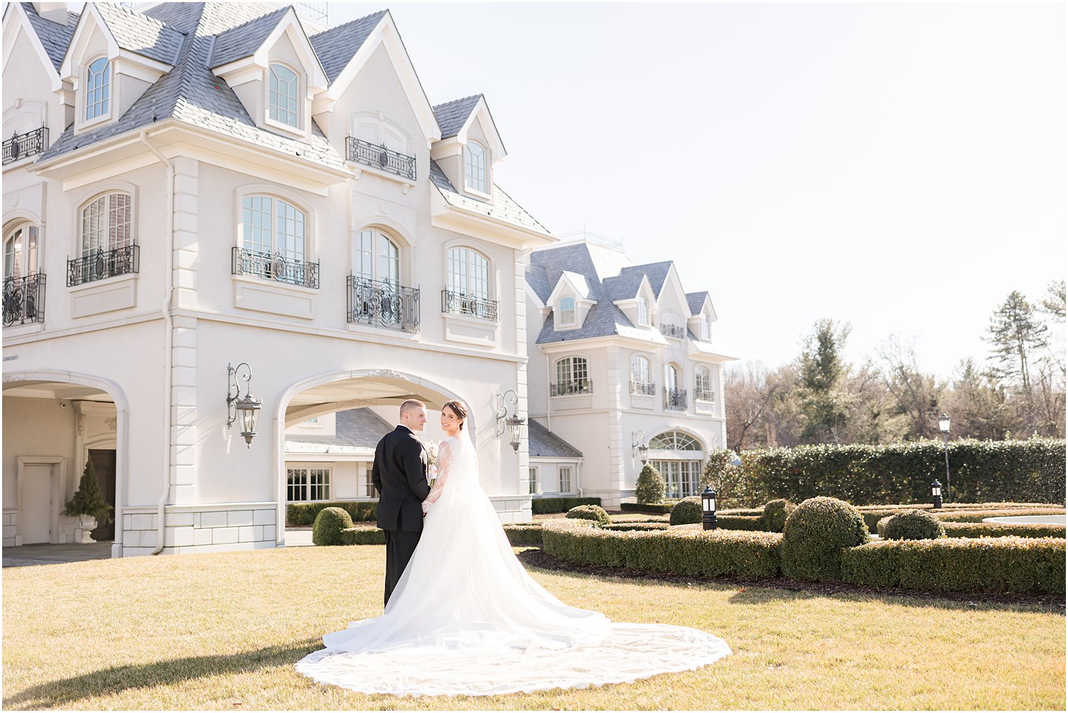newlyweds stand on lawn in front of Park Chateau Estate with bride's train spread out
