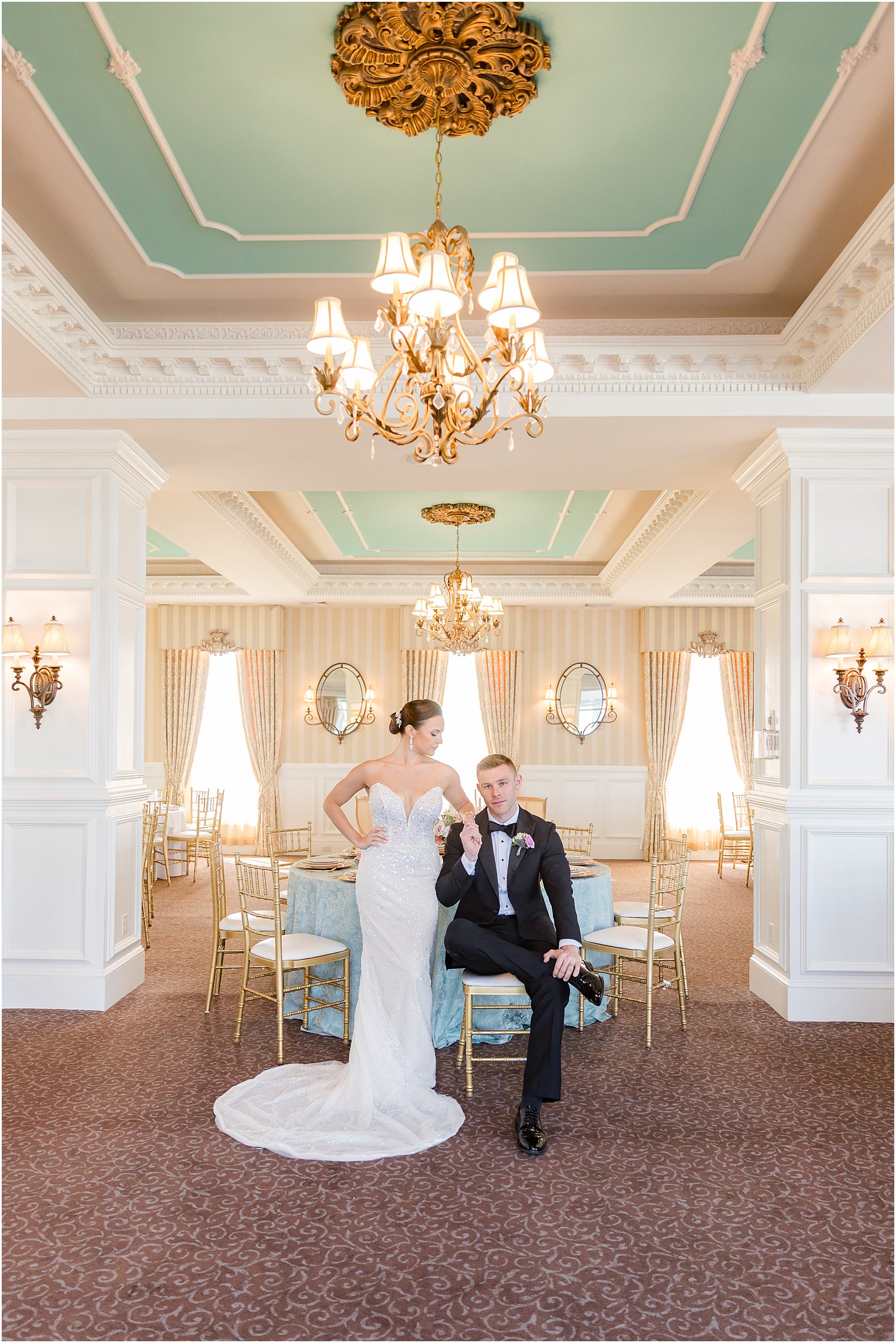 groom sits on blue chair inside Mallard Island Yacht Club ballroom with bride leaning on his shoulder