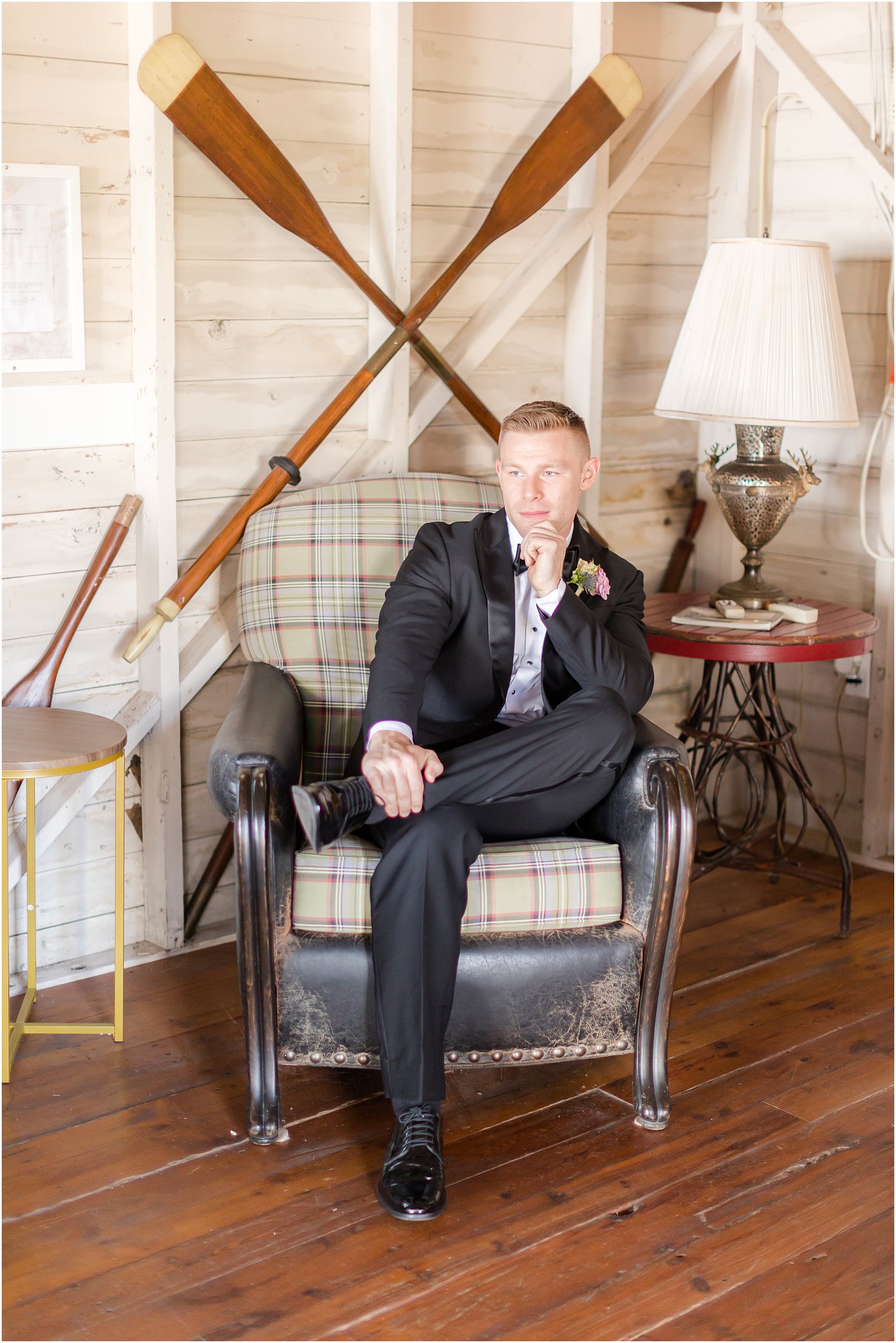 groom sits in chair in front of oars at Mallard Island Yacht Club