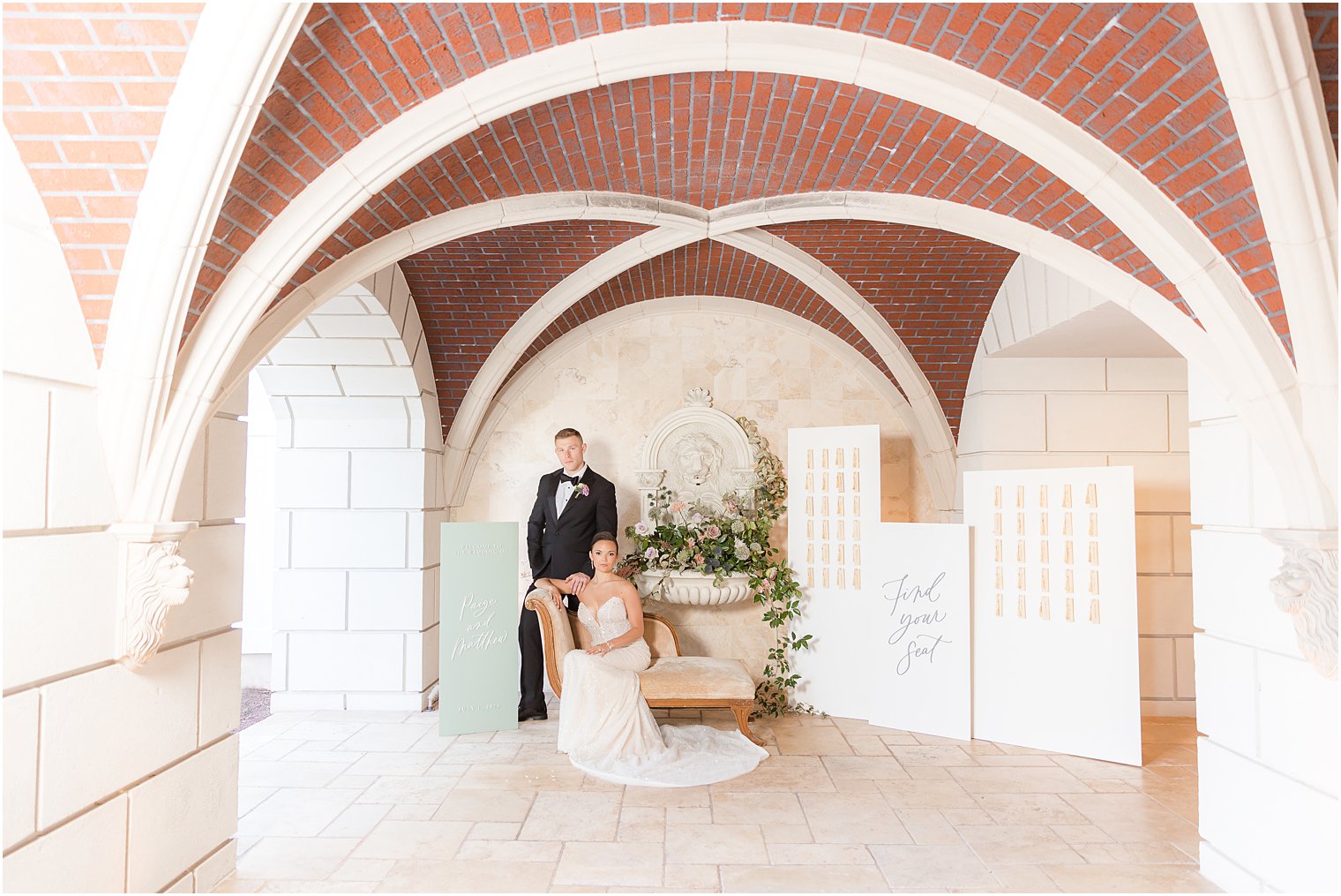 bride sits in chair while groom stands beside it between seating charts