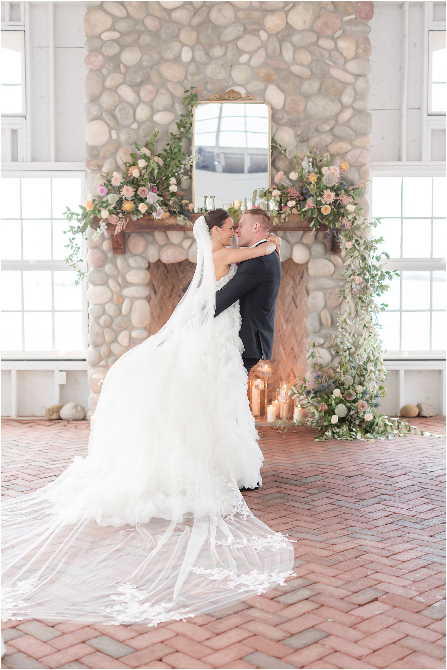 groom lifts up bride standing inside barn at Mallard Island Yacht Club with stone fireplace behind them