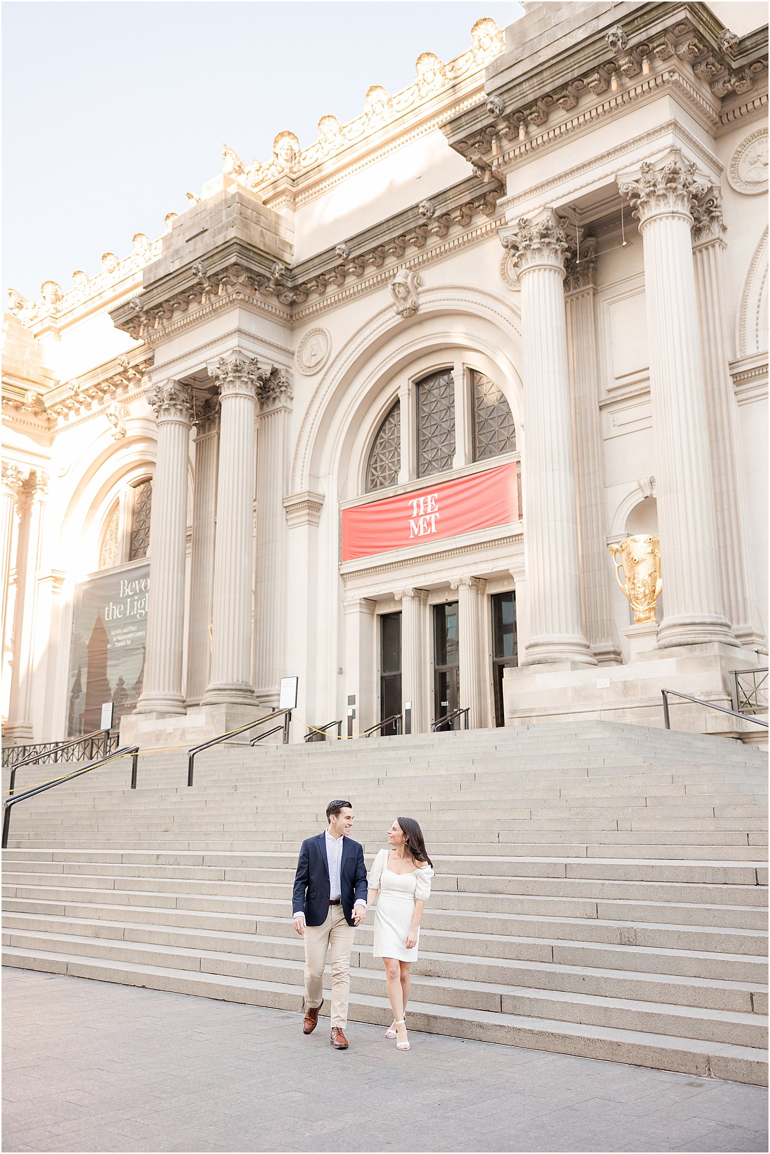 groom in navy blazer helps bride in white dress down steps of the MET Museum in NYC