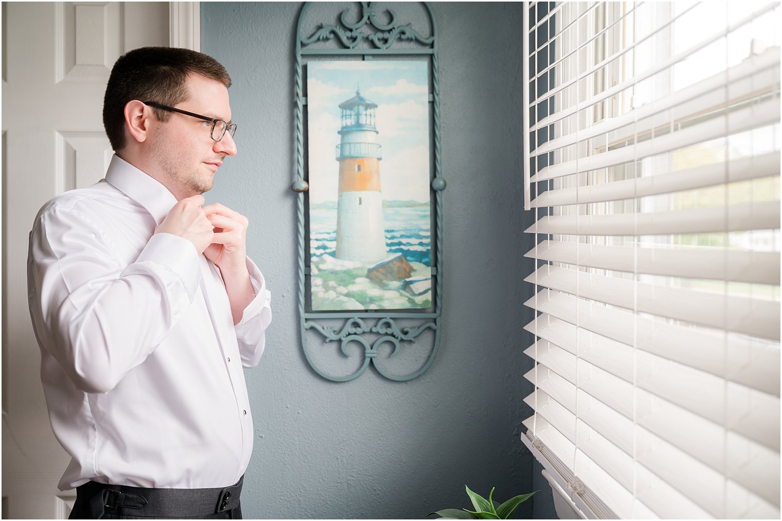 groom preparers for wedding day looking out window with lighthouse painting behind him