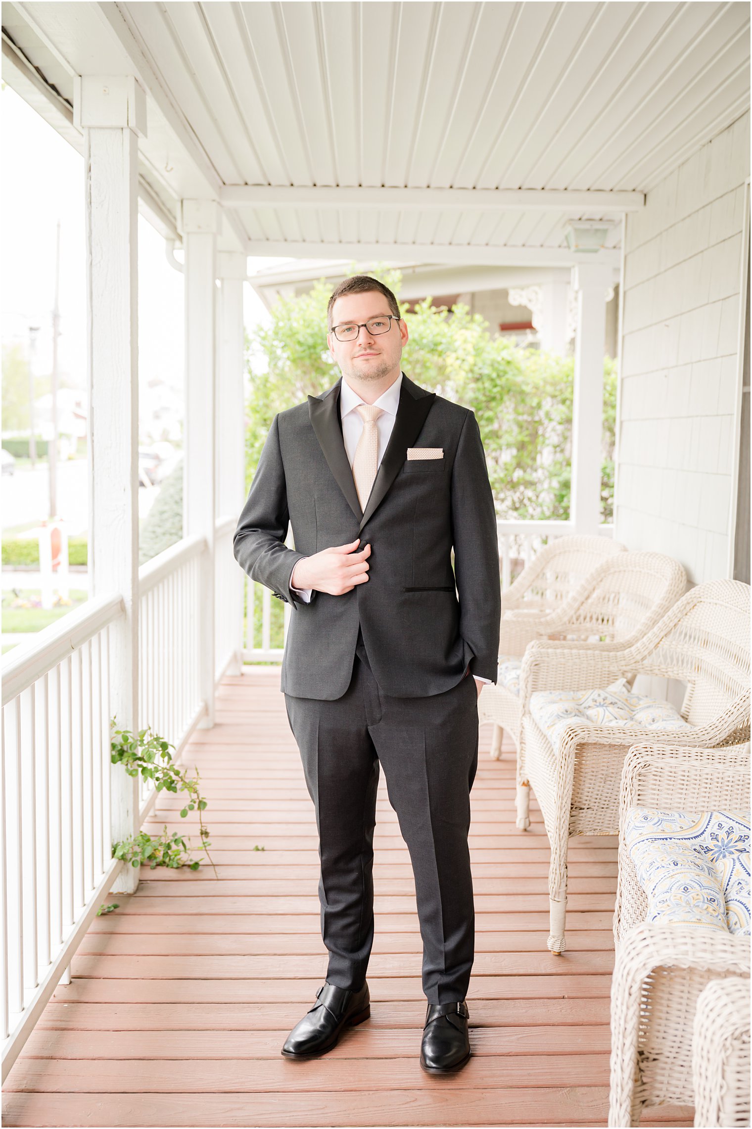 groom stands on front porch holding lapel of suit jacket 