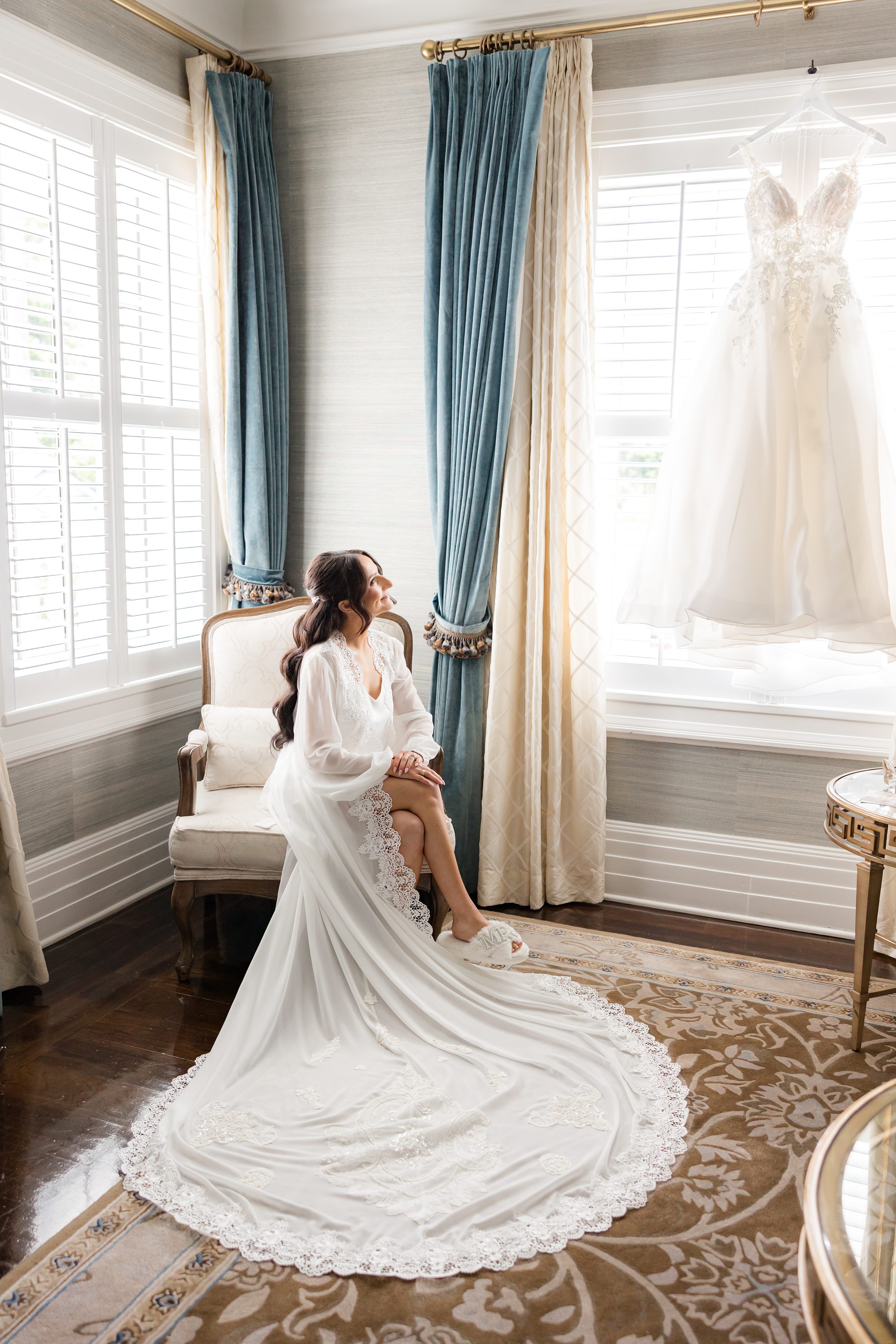 Bride wearing a lace white gown while looking at her lace wedding gown hanging in the window