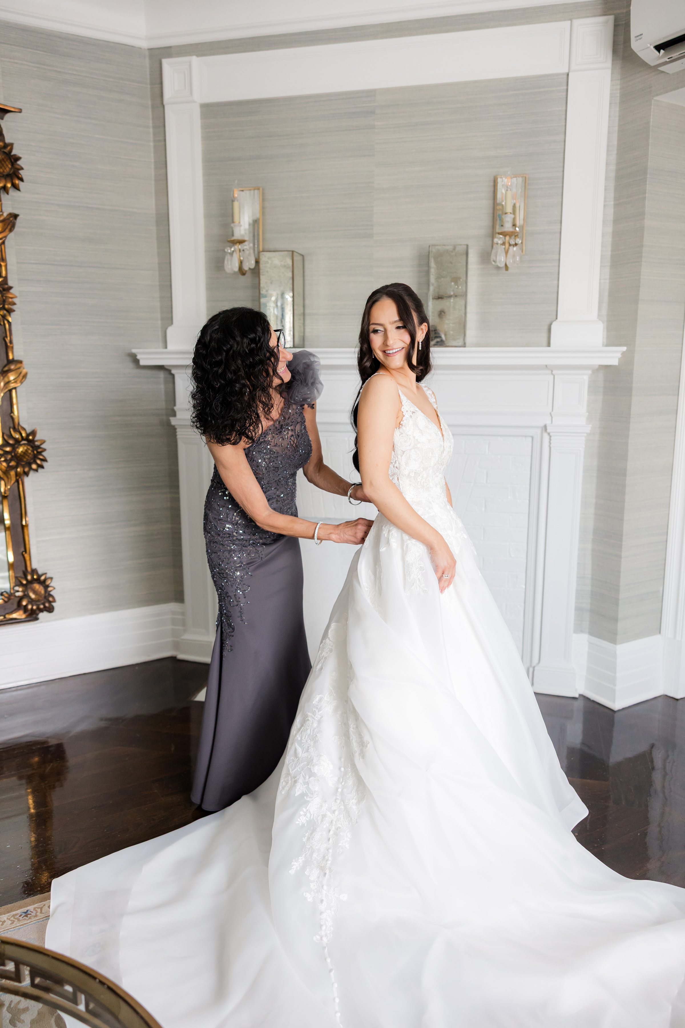 Bride's mother is helping the bride wear the gown as they smile at each other