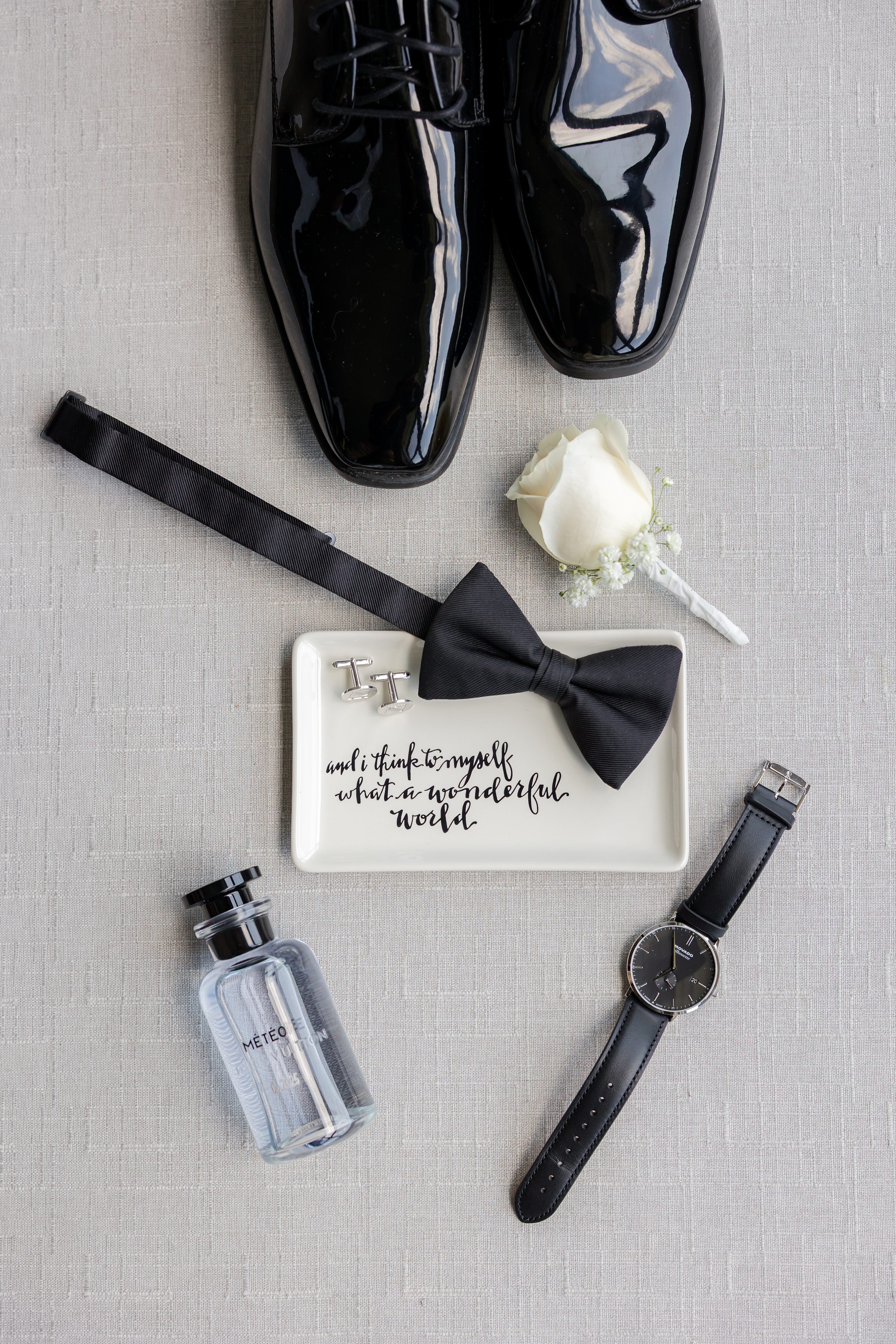 Groom's close-up picture of black watch, perfume, shoes , ribbon and white flower