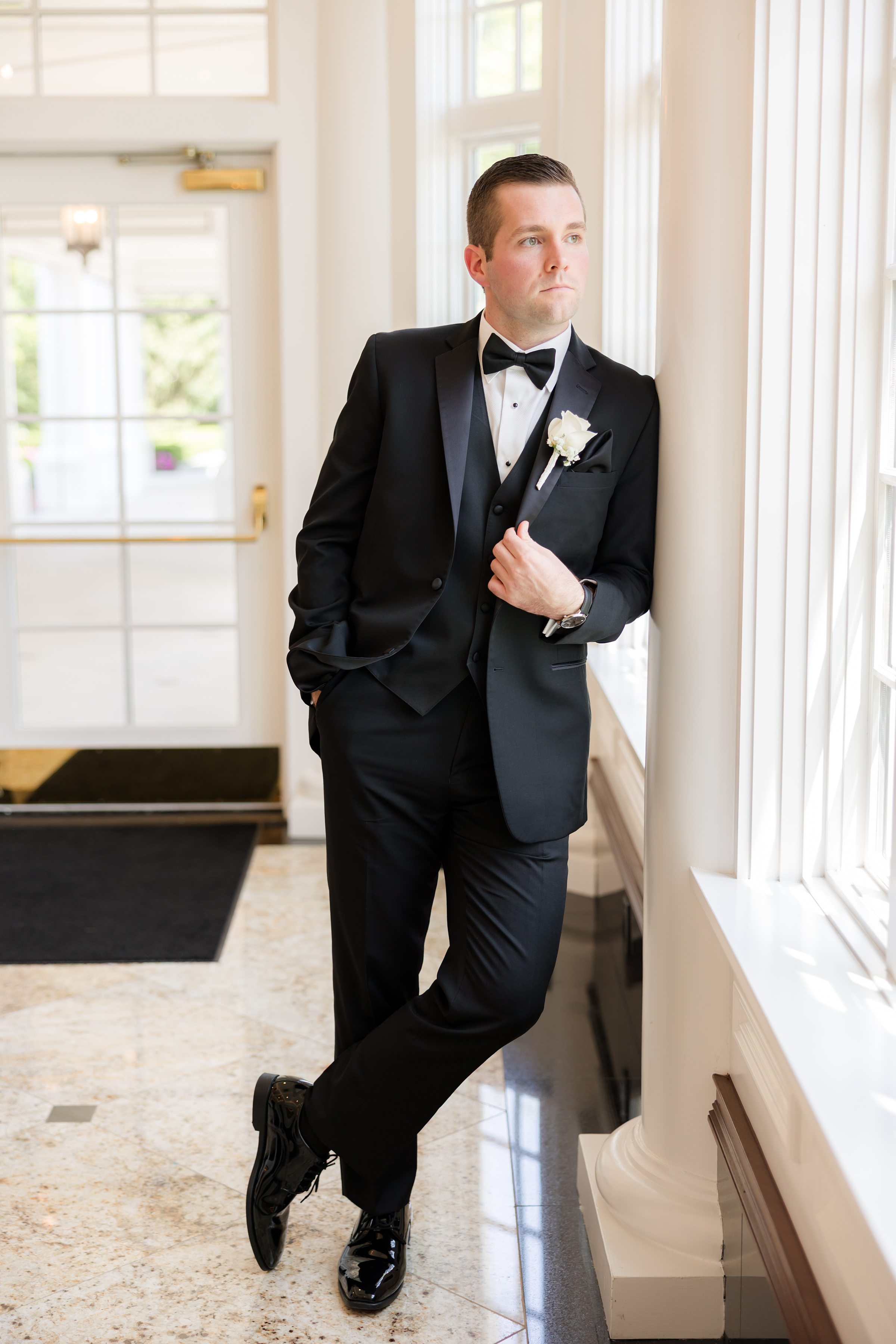 A solo shot of groom wearing black tuxedo, standing looking to the window 