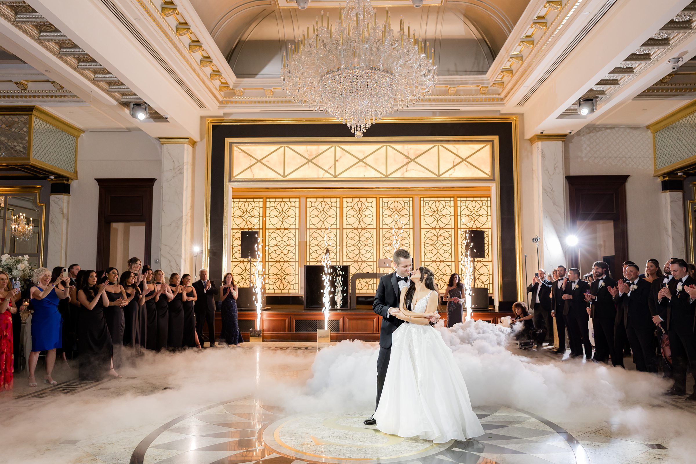 The couple are dancing in front of the guests with mini fireworks at the back and some smoke on the floor 