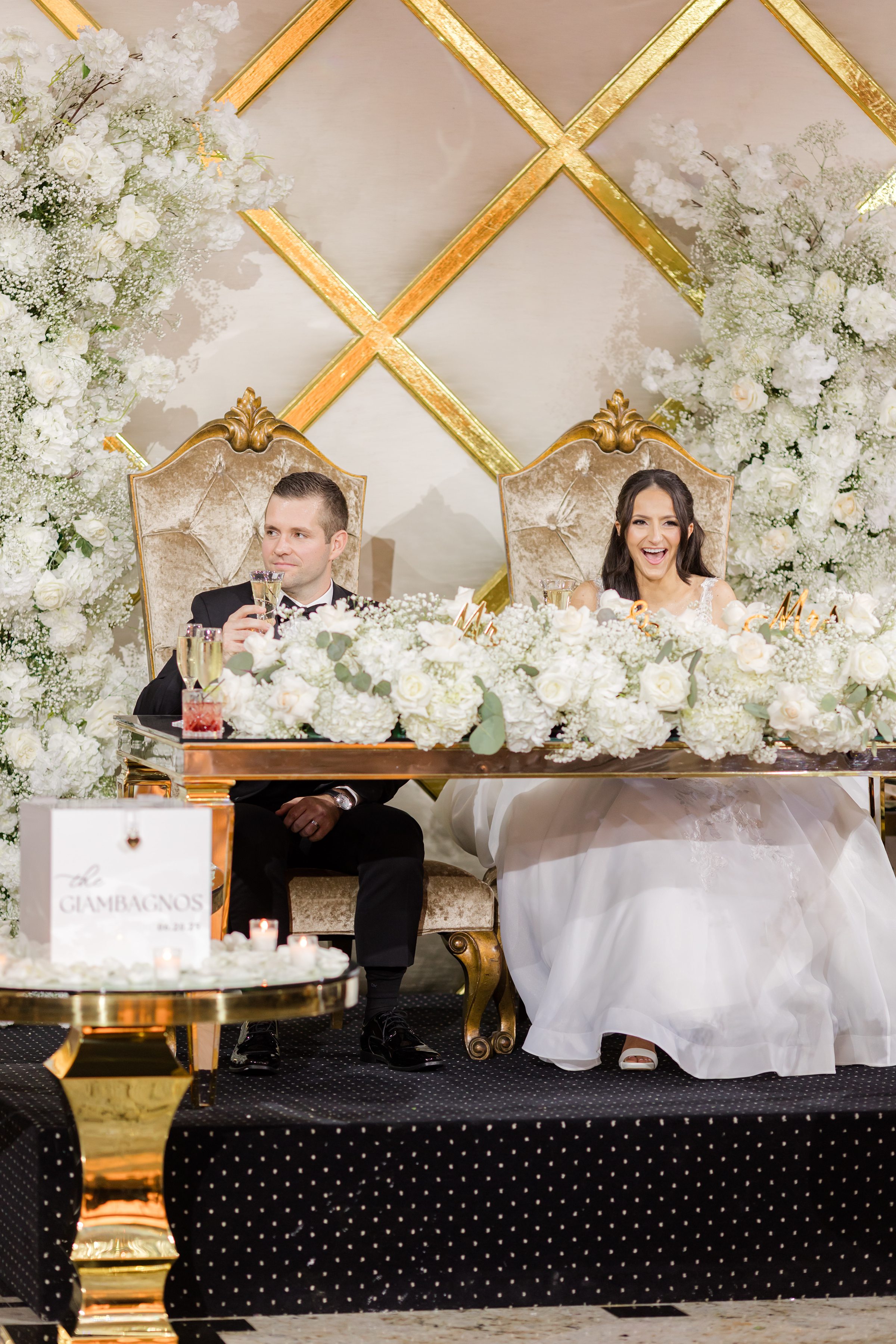 The couple are sitting with lots of flowers on the table and the groom is holding a glass of champagne 