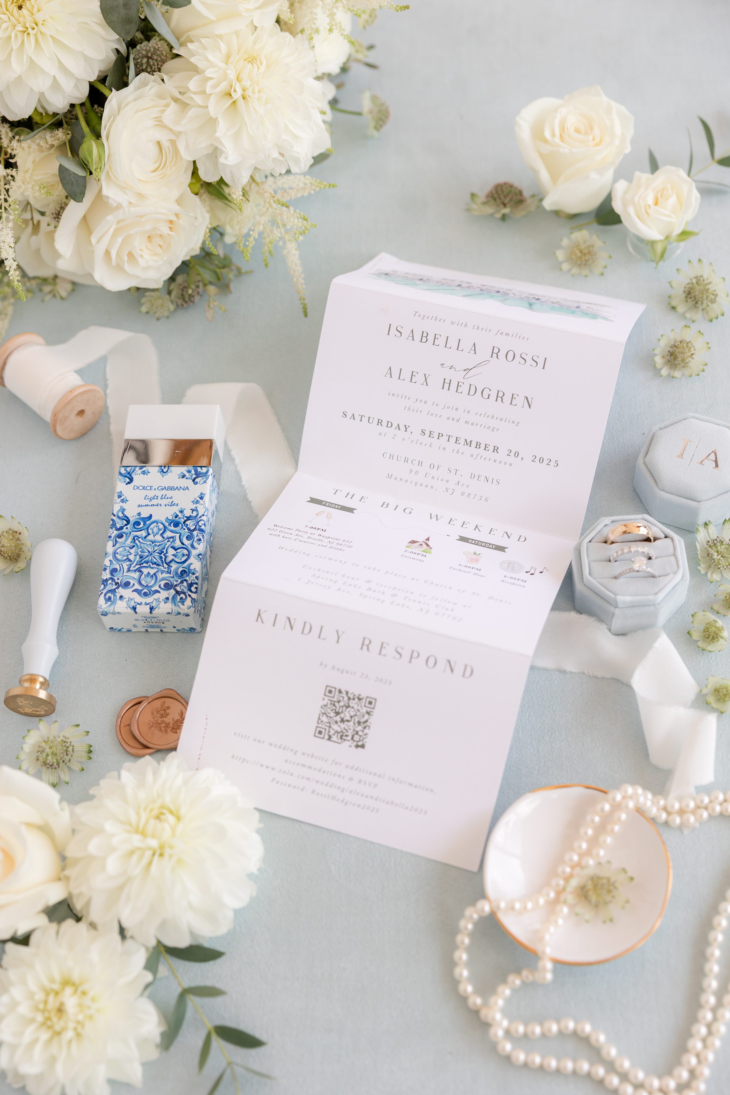 Wedding invitation perfume, pearl necklace, ribbon, and floral accents arranged on a soft blue surface.