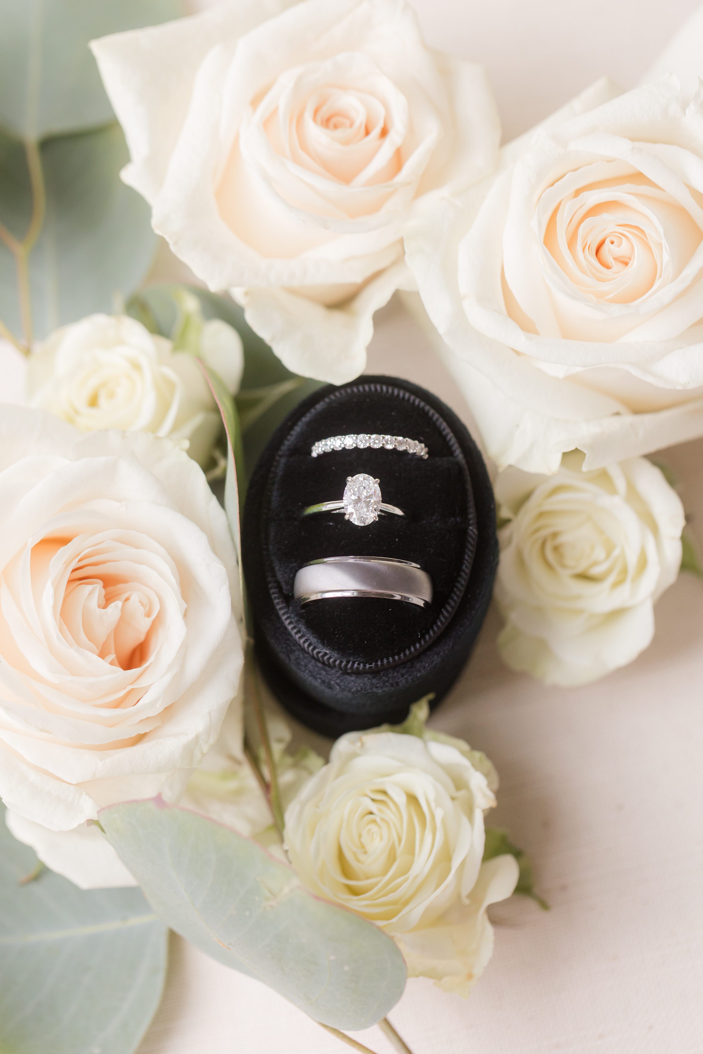 Photo of silver engagement ring and wedding bands in a black ring box, surrounded by white roses