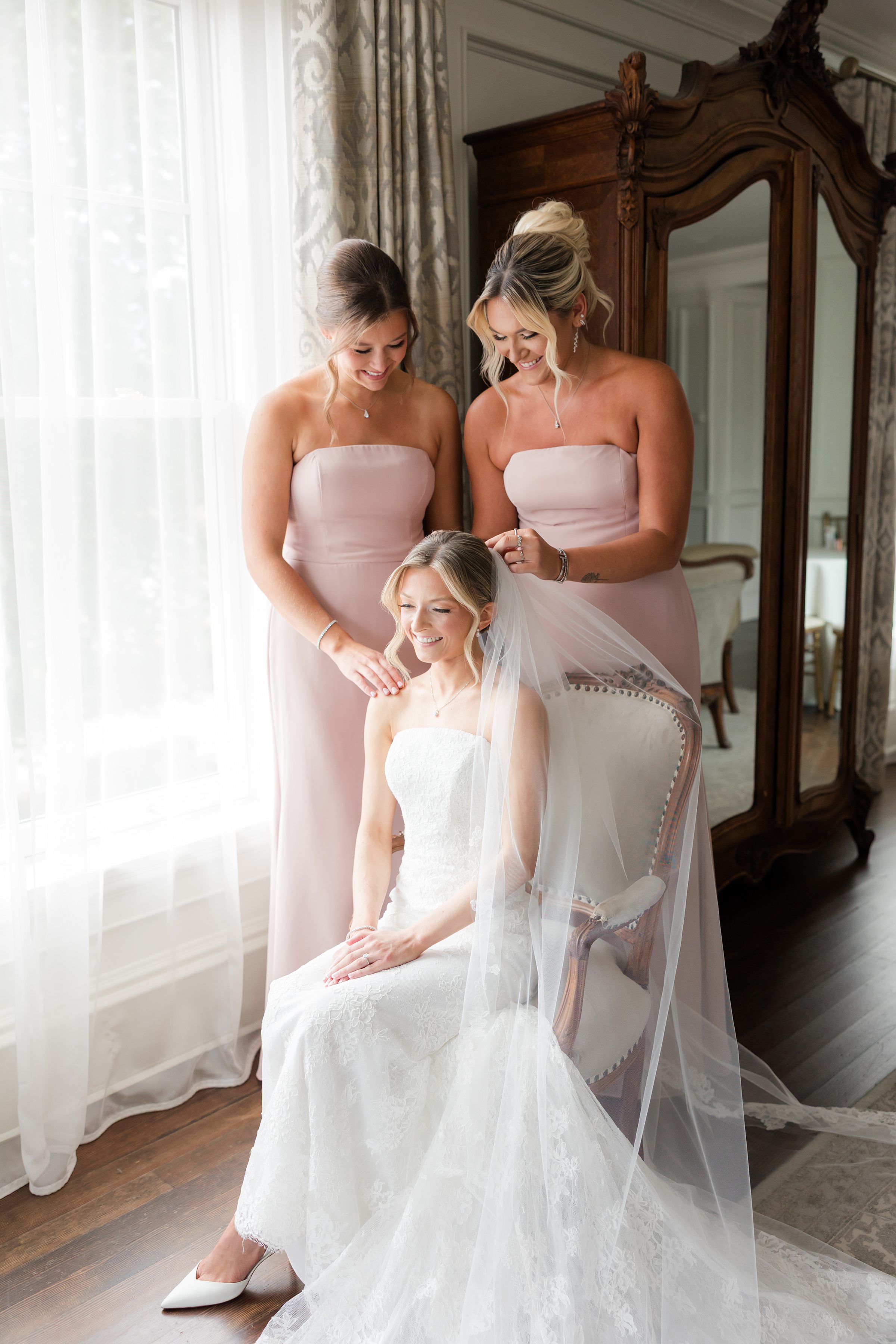 Bride seated on a wooden-cushioned chair while 2 of her bridesmaids are putting her veil on