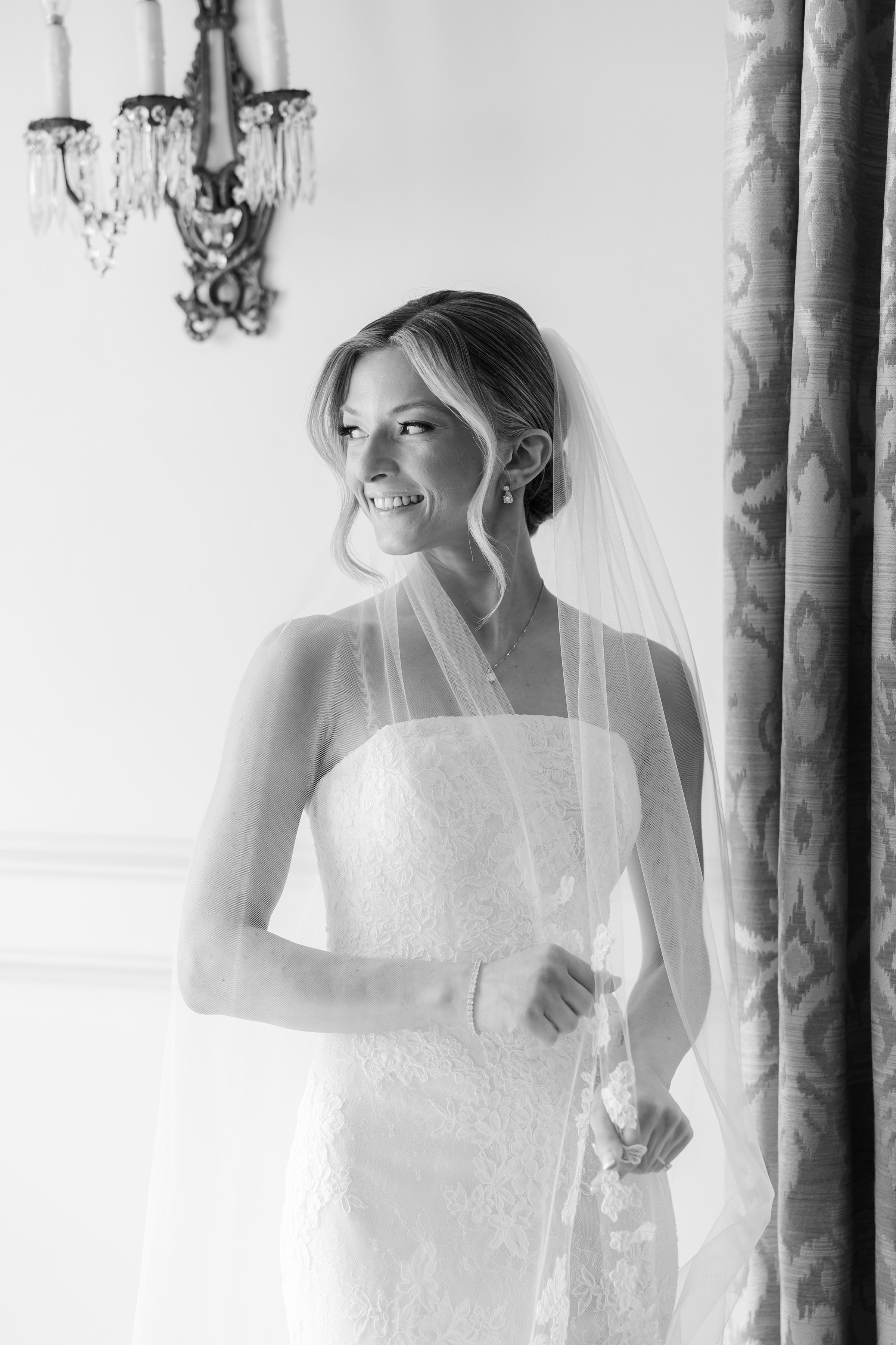 Black and white half-body photo of bride holding her veil while looking out the window