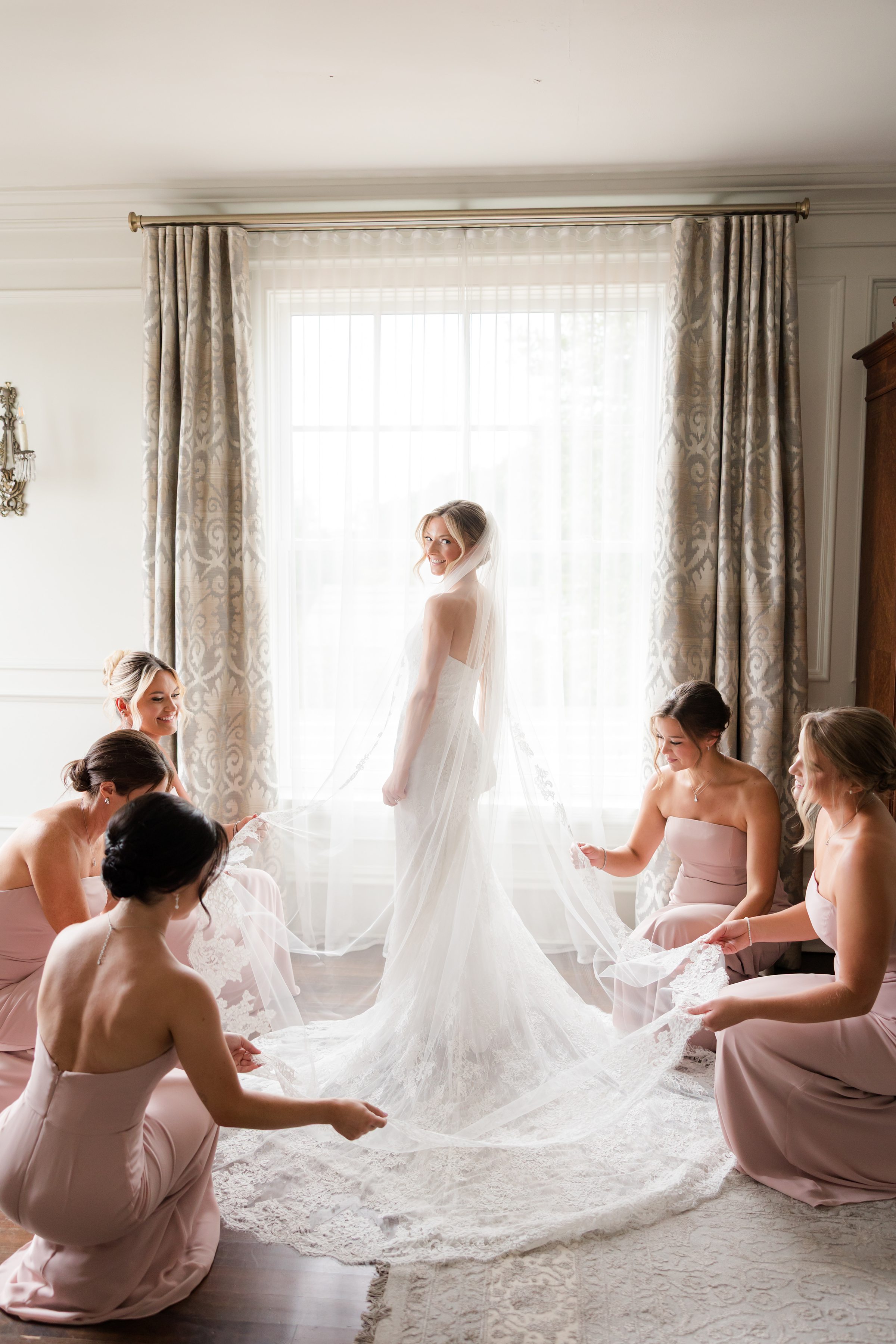 Photo of bride and her bridesmaids by the window. Bridesmaids are helping fix the bride's veil while she looks back to the camera