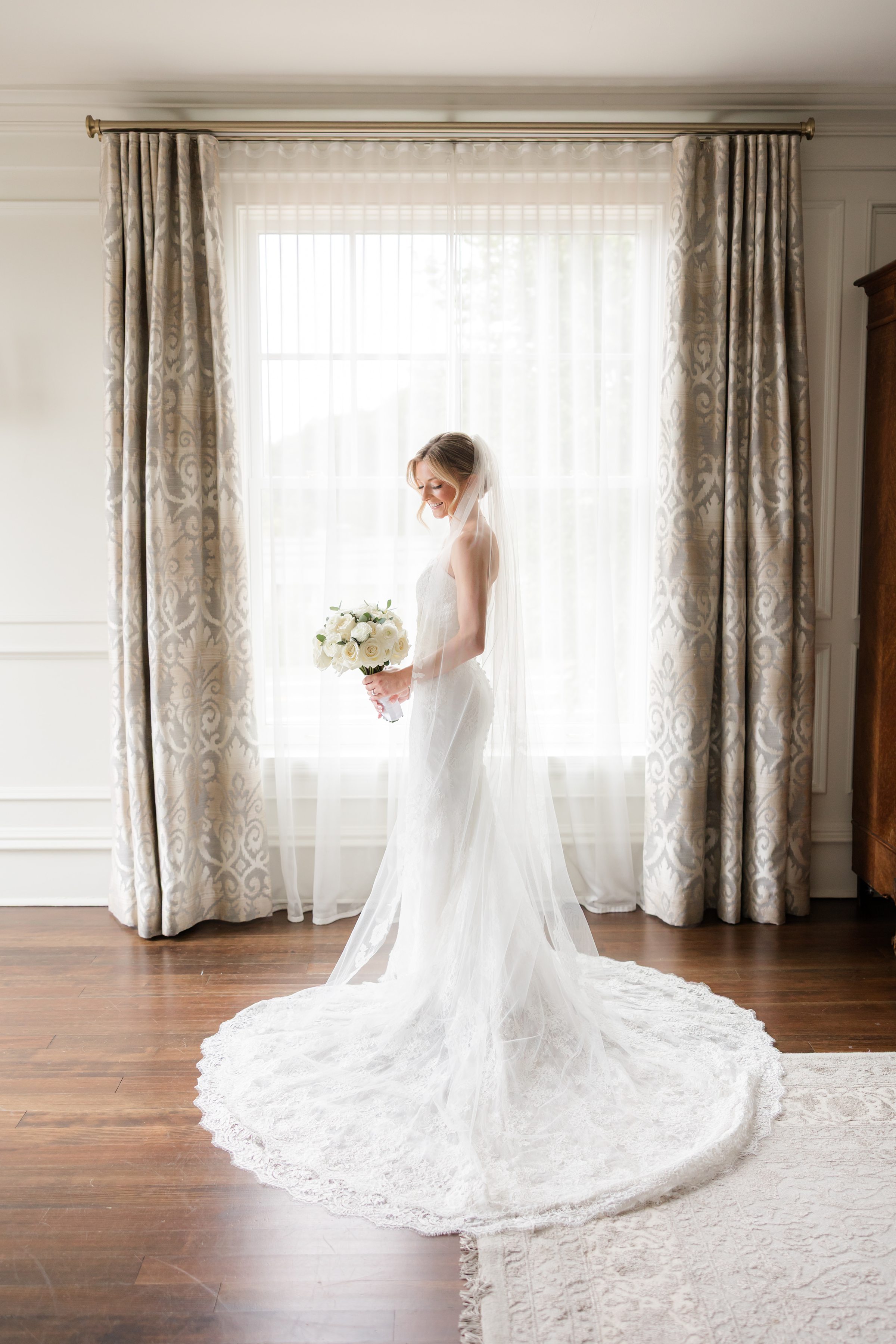 Bride standing sideways, looking at her bouquet, by the window with sheer curtains