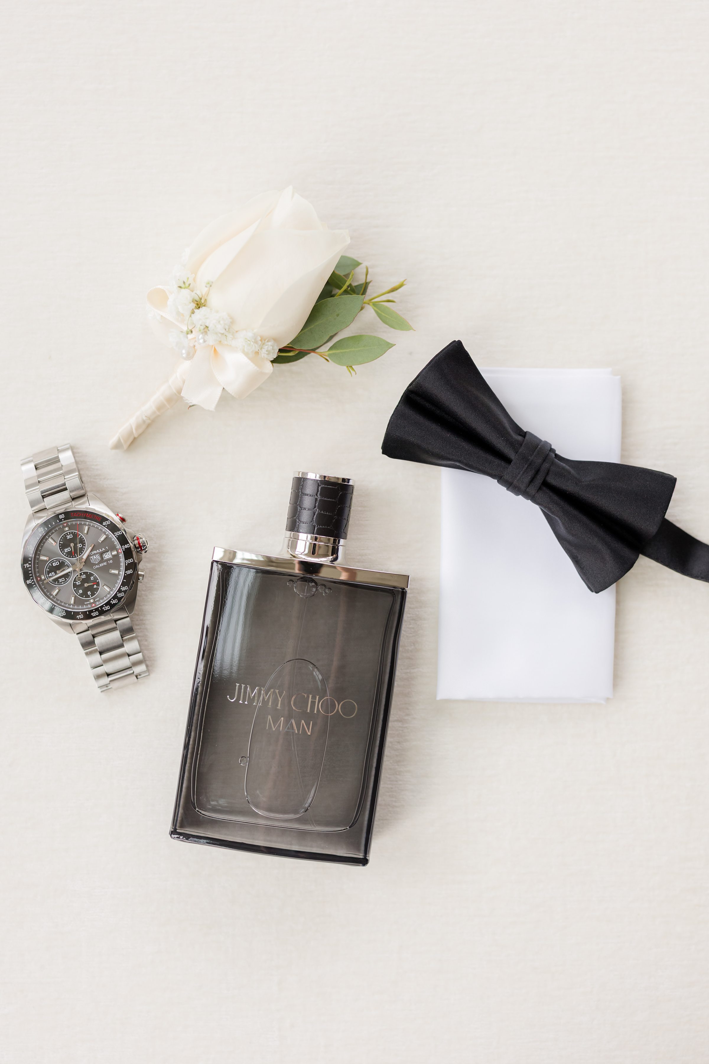 Detail shot of groom's items, including a silver watch with black accents, black Jimmy Choo perfume, black bow-tie, and a white rose corsage