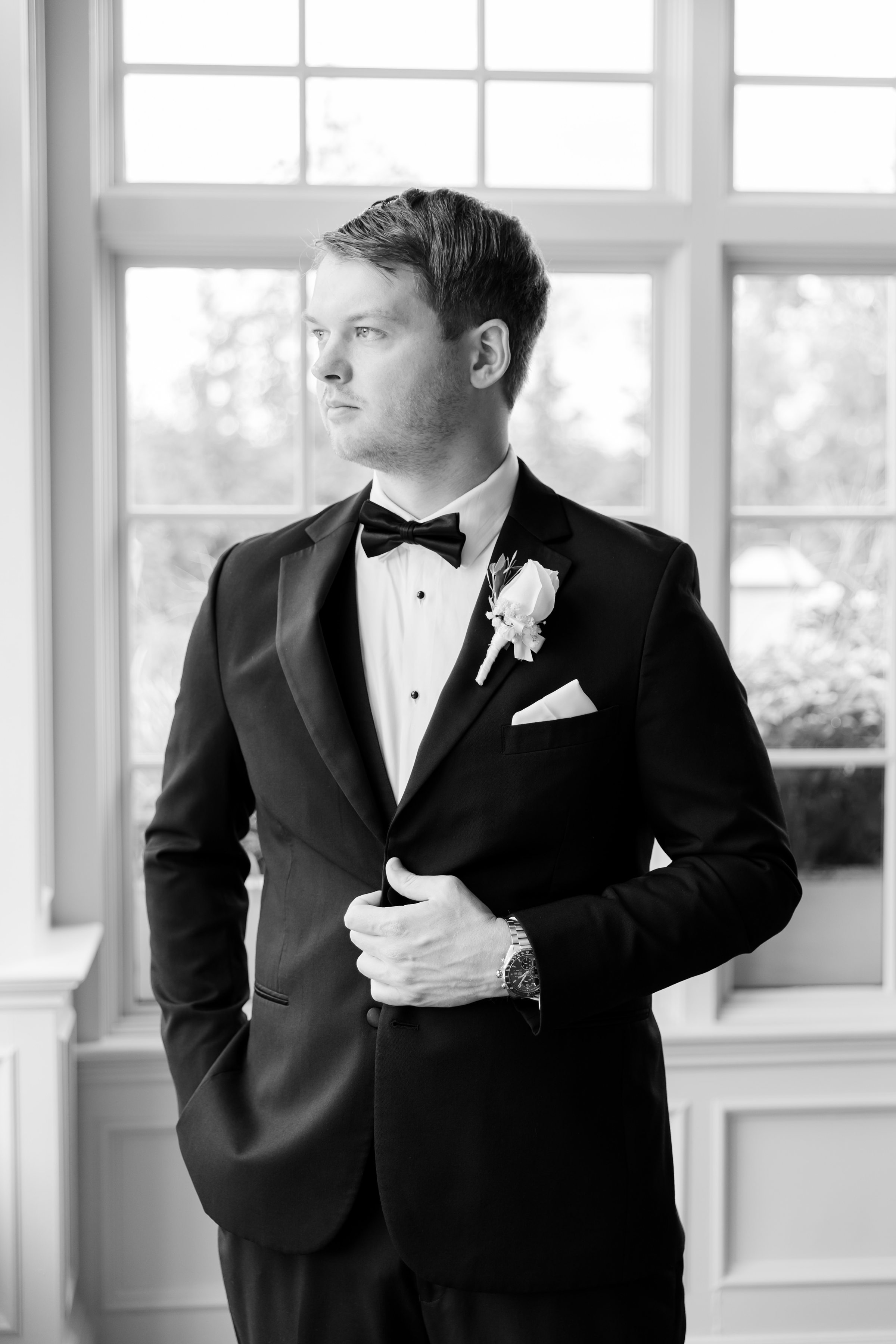 Black and White portrait of the groom looking out the window while his left arm is holding his suit, and right arm inside his pocket