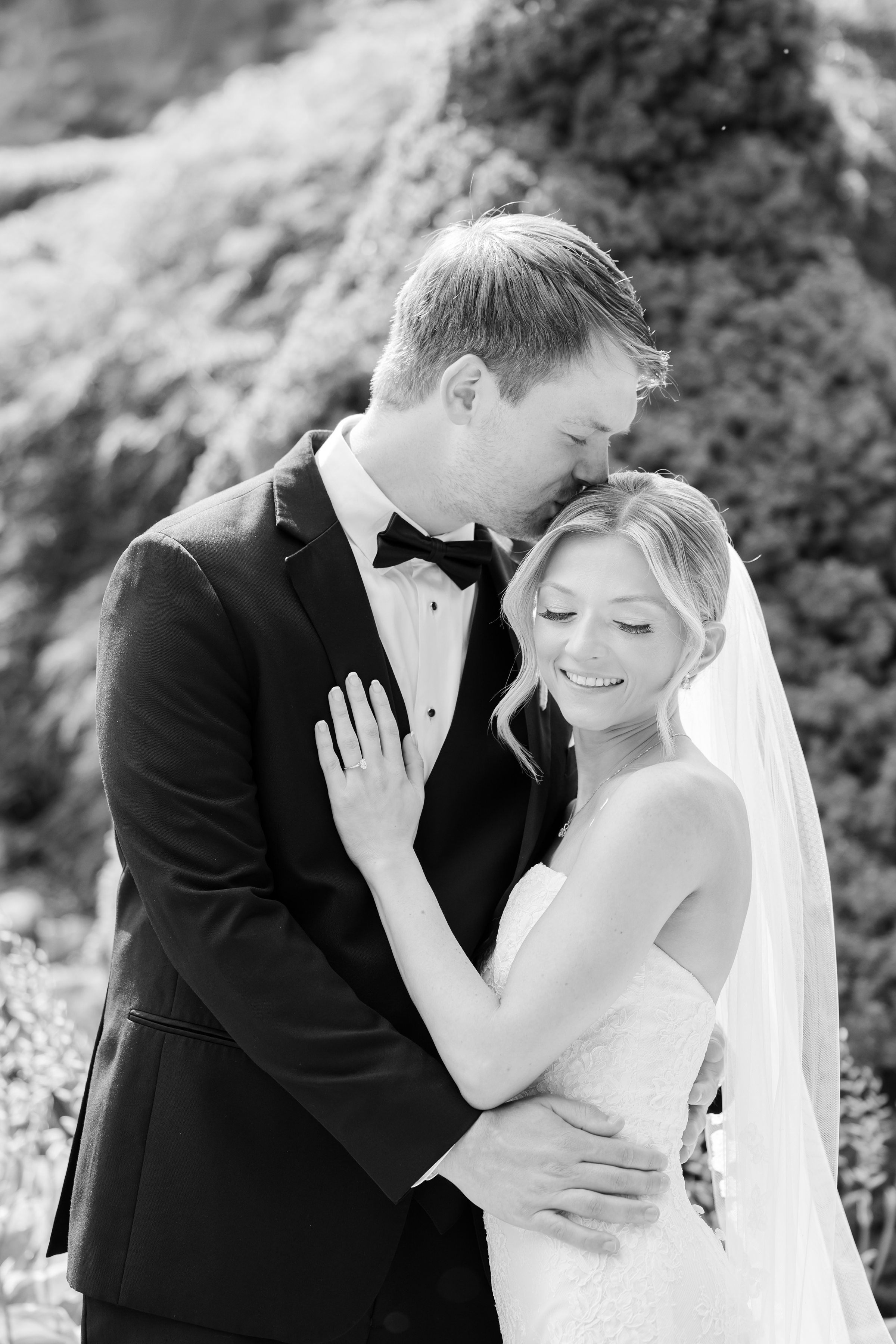 Black and white portrait of the groom kissing the bride's head while his hand is wrapped around her waist, and her touching his chest with her eyes closed