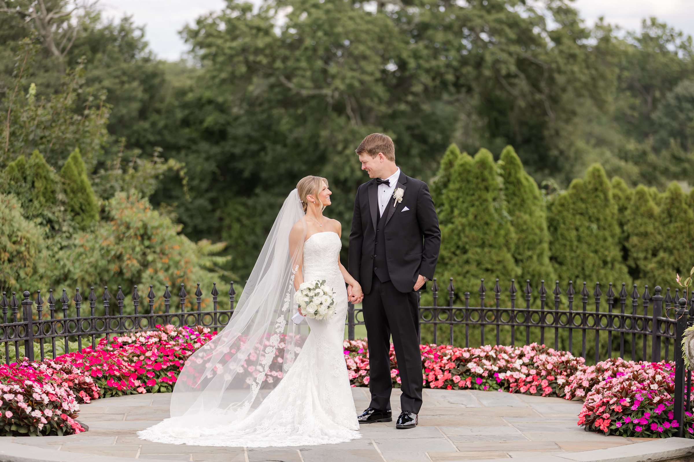 Landscape photo of the bride and groom looking at each other in the middle of a veranda filled with pink flowers