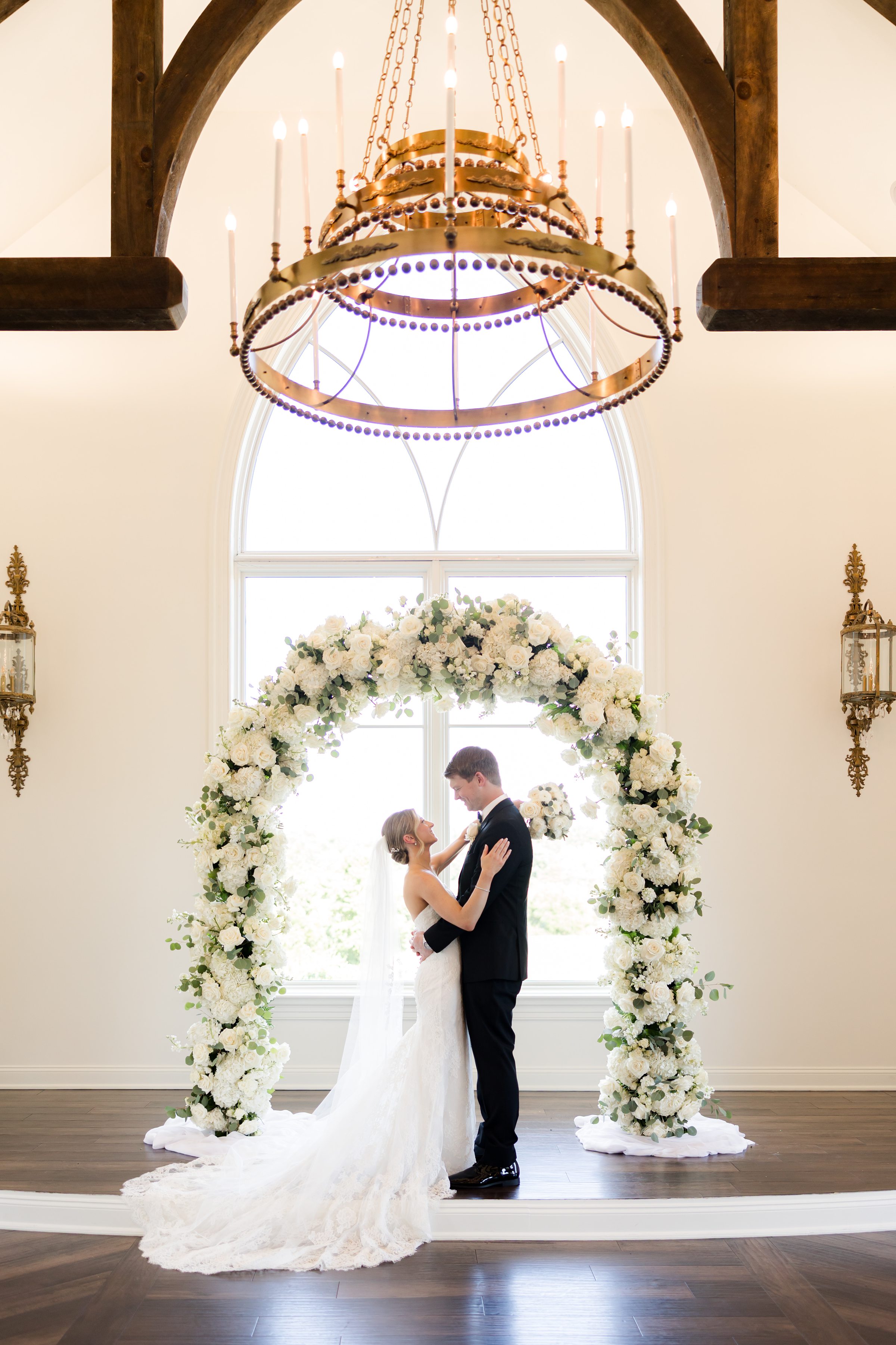 Bride and groom holding and looking at each other by the arc, under a golden chandelier, in the chapel