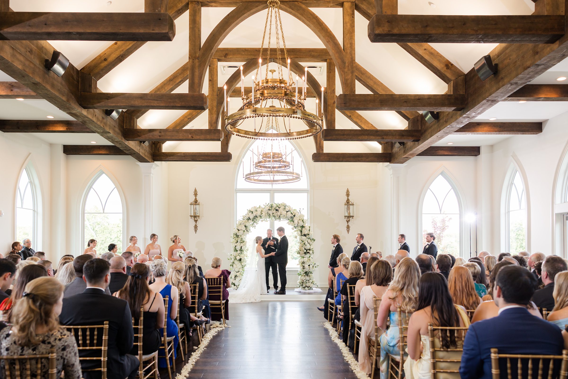 Wide shot of a filled ceremony space, with groom, bride, and officiant at the center, and the wedding party to the side