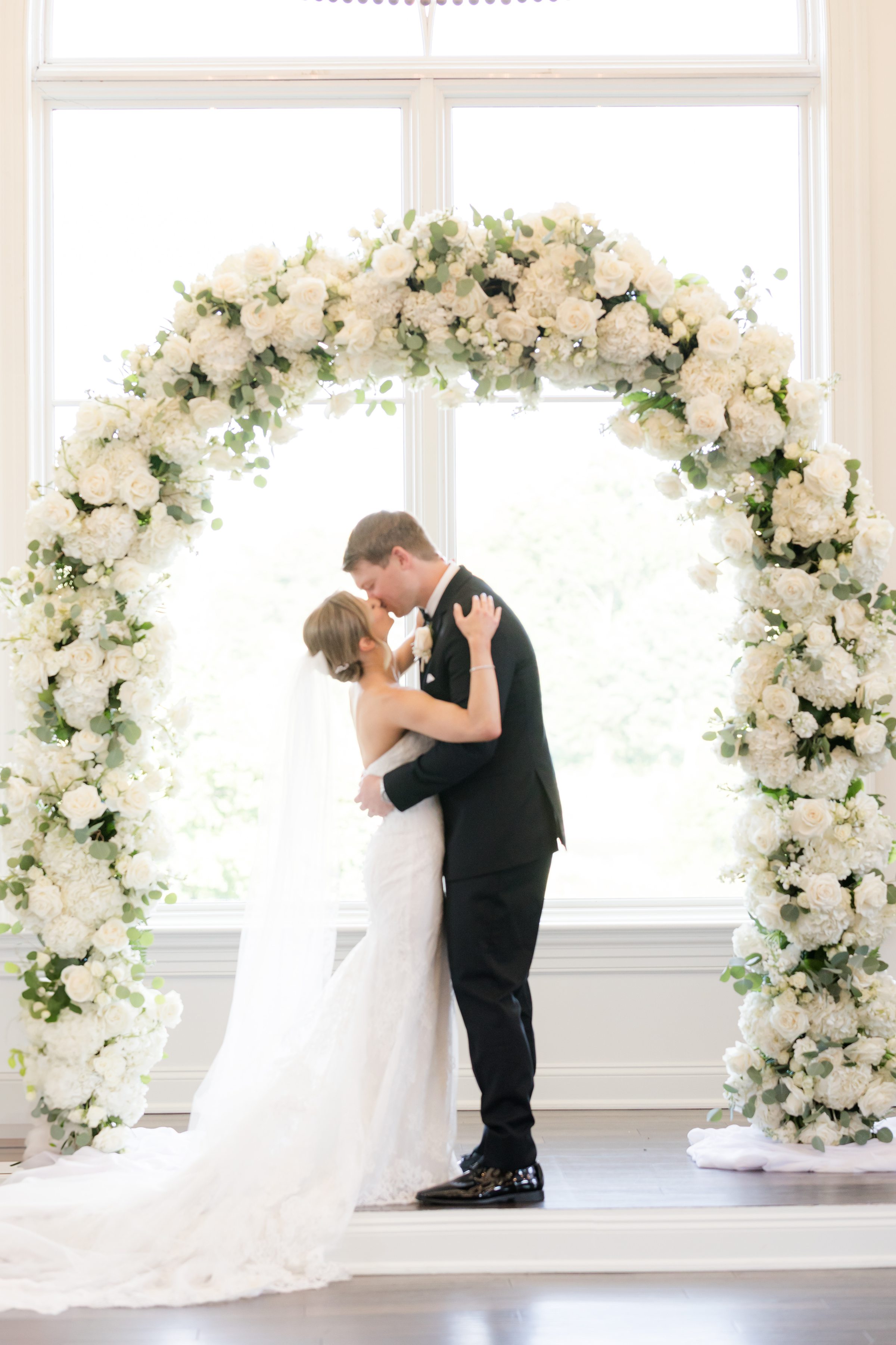 Bride and groom first kiss as husband and wife, by the white flower arch