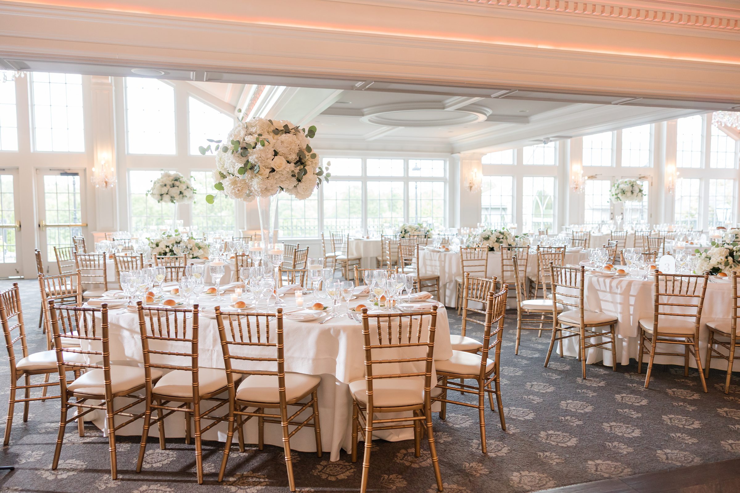An empty, decorated ballroom, with wooden chairs and white cushion. Glass vase filled with white flower as table toppers