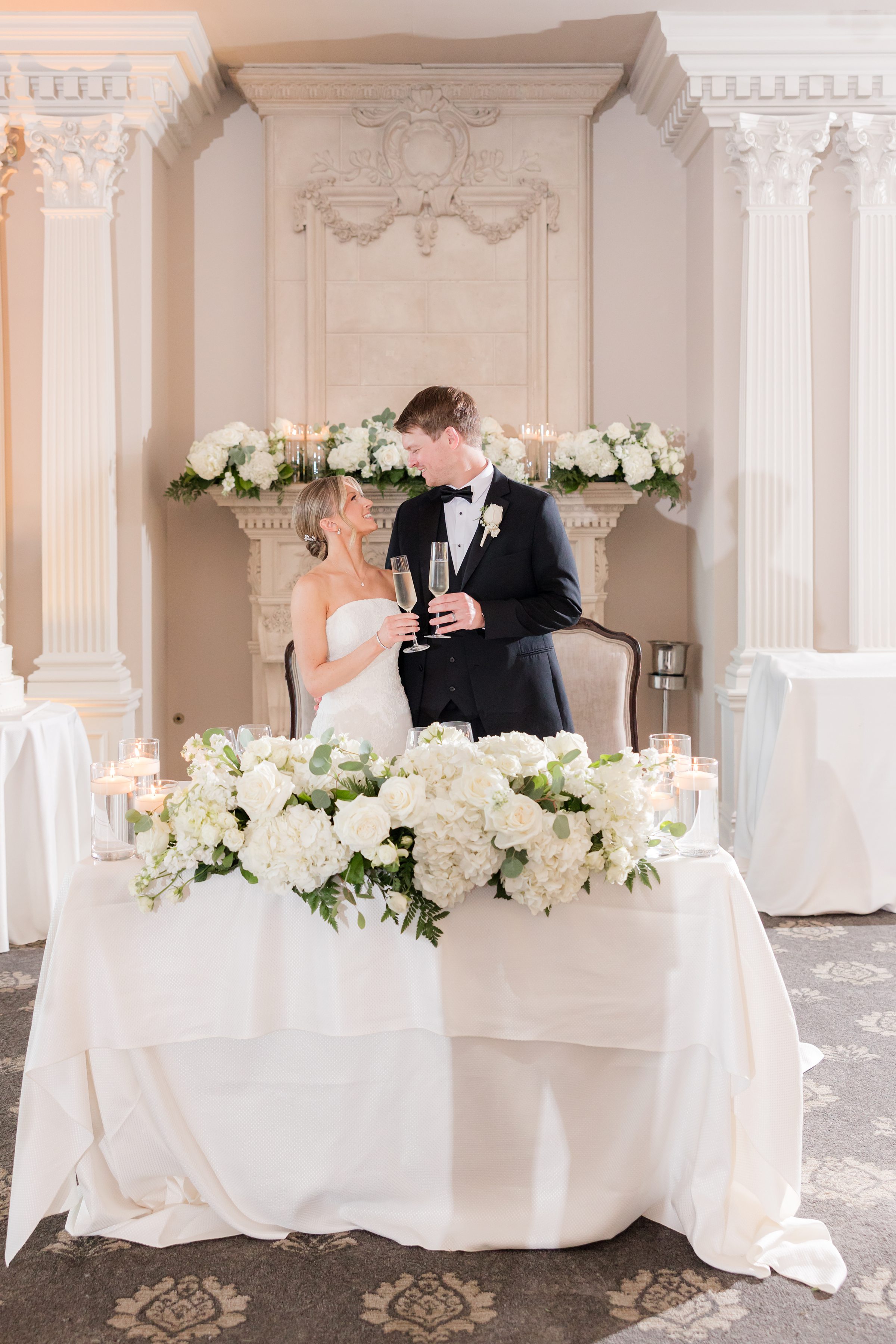 Photo of bride and groom standing by the sweetheart table, holding a glass of champagne in each of their hands while looking at each other