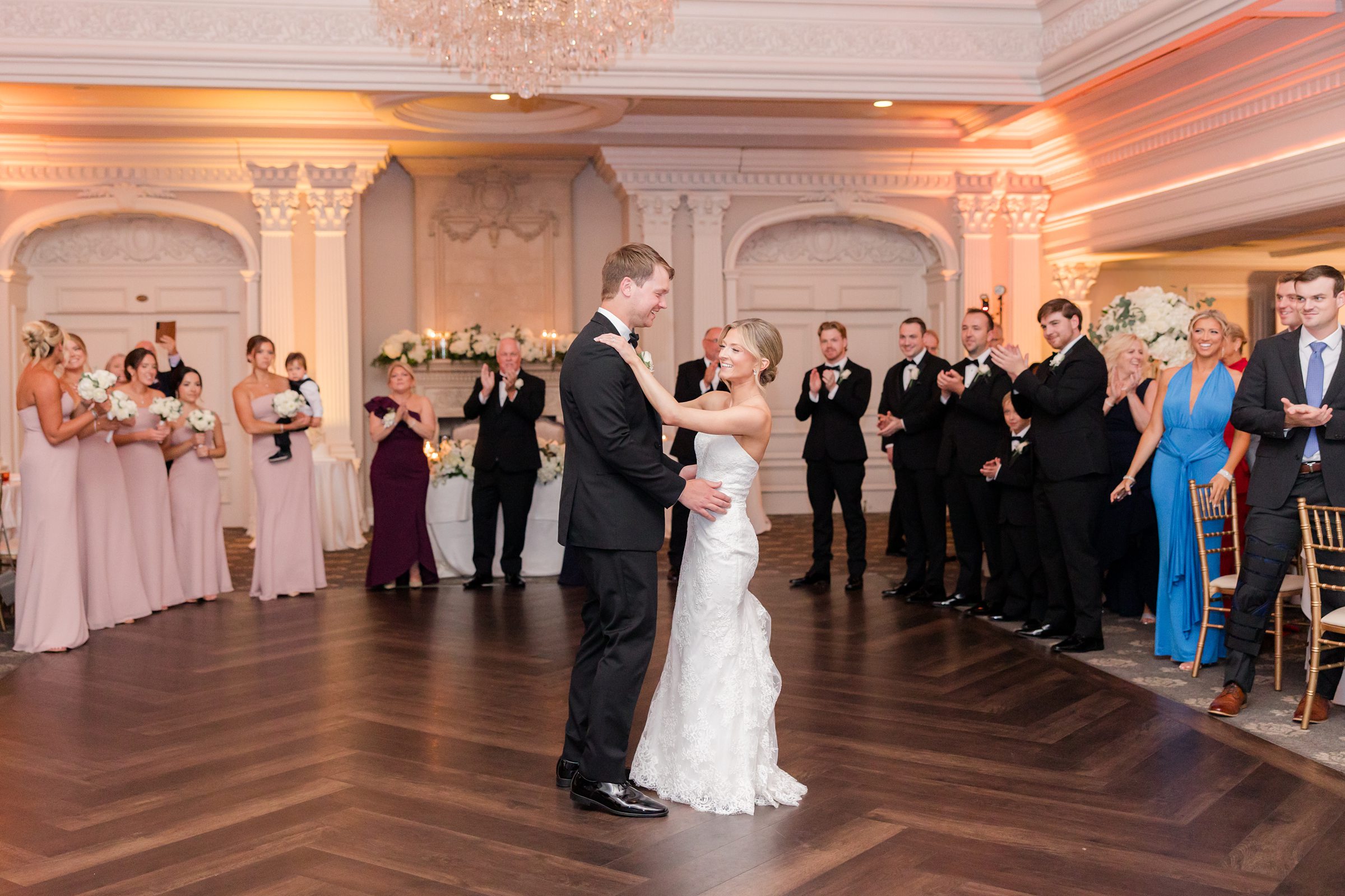 Bride and groom dancing on a wooden dance floor, holding each other while surrounded by guests