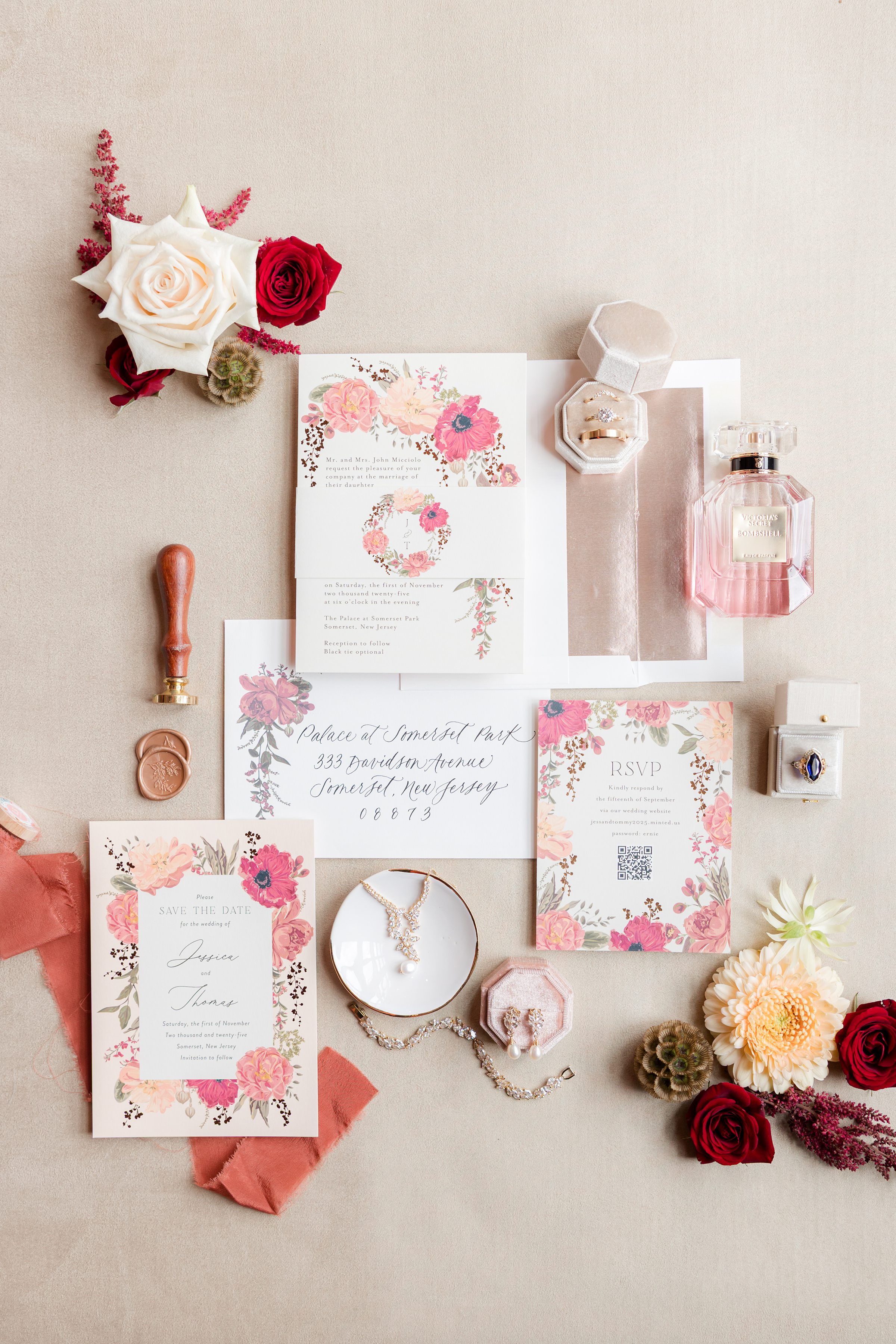 Wedding invitation flat lay suite adorned with soft blush and coral florals, featuring delicate stationery, rings in a velvet box, a wax seal, perfume, and elegant jewelry, beautifully styled to evoke a timeless love story.