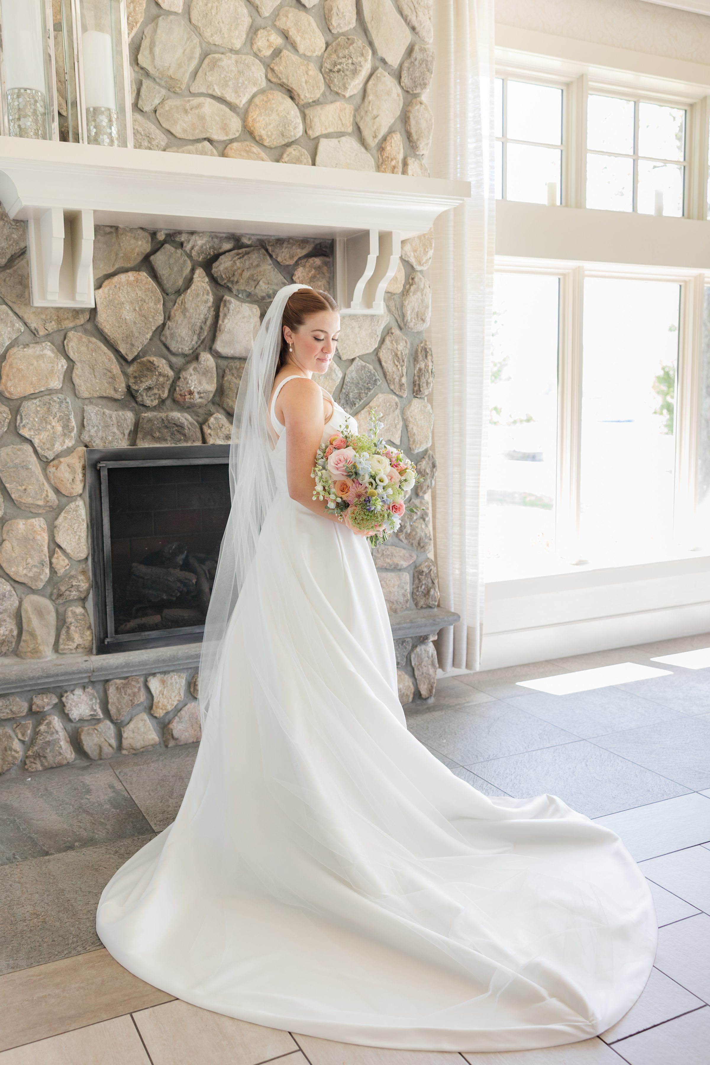 full body portrait of bride wearing plain white wedding dress with a full circular train and a long veil holding a bouquet with pink, orange, white flowers 
