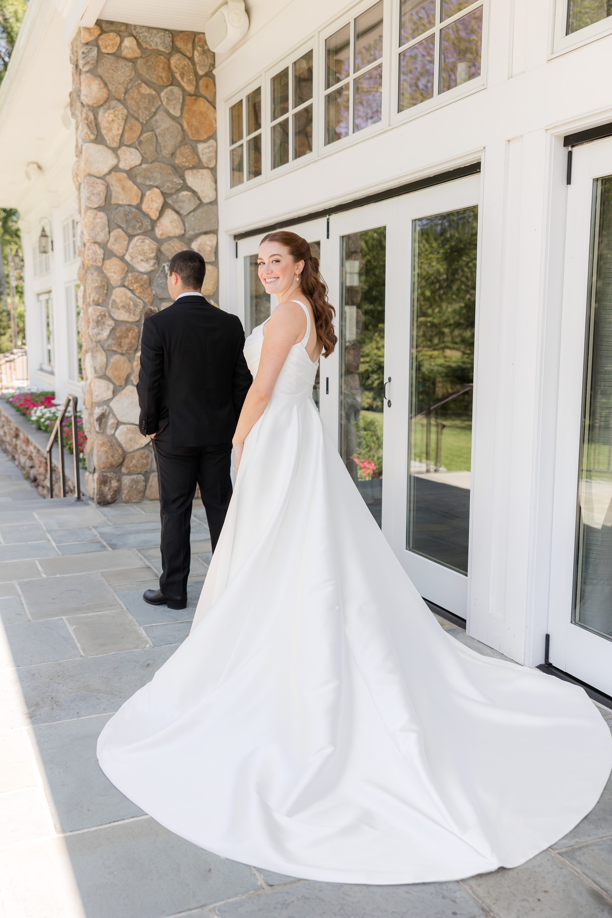 A portrait picture of bride standing, smiling, looking back for the camera while the groom is turning his back