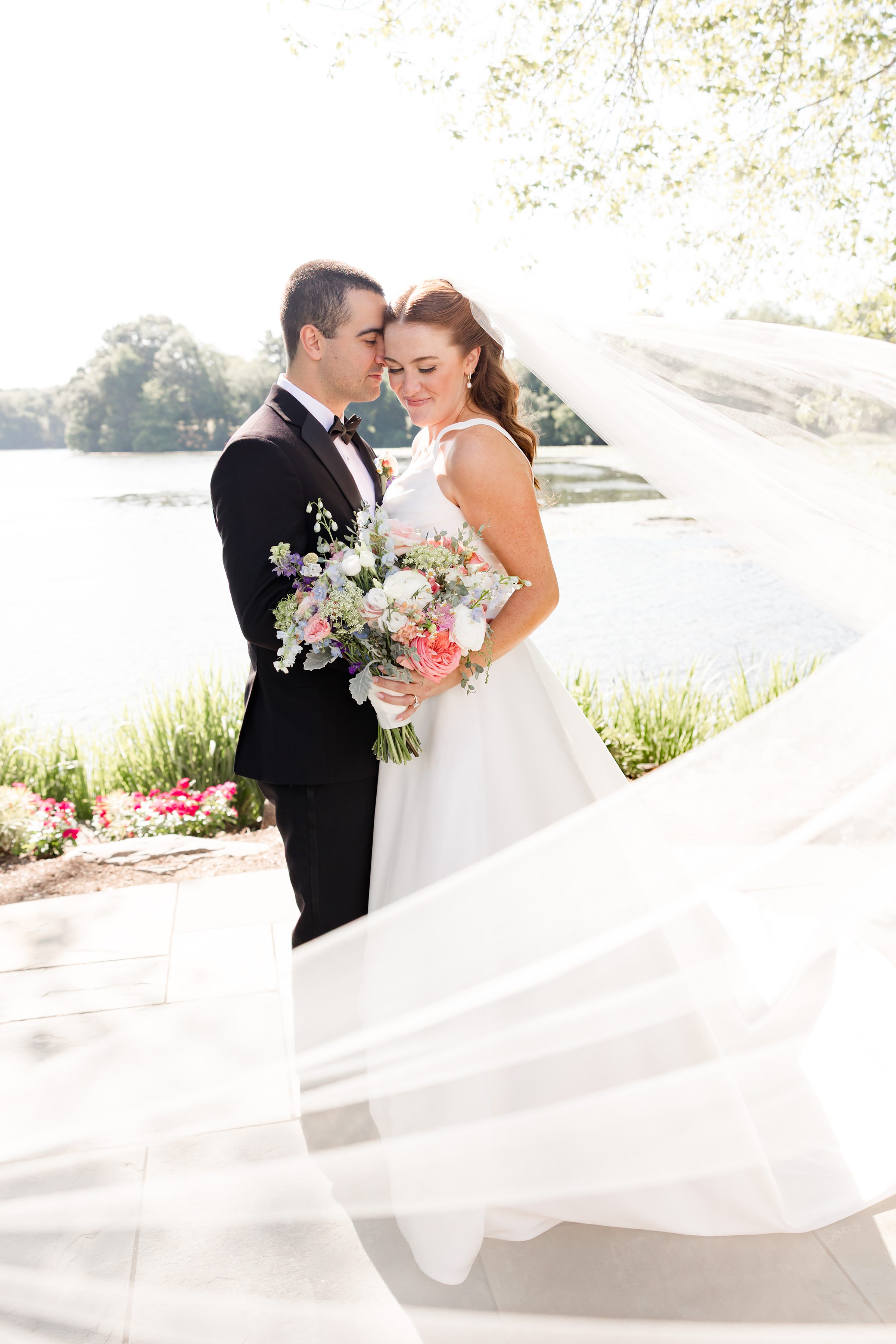 portrait of bride and groom being close to each other while bride is holding the wedding bouquet with veil blowing to the side 