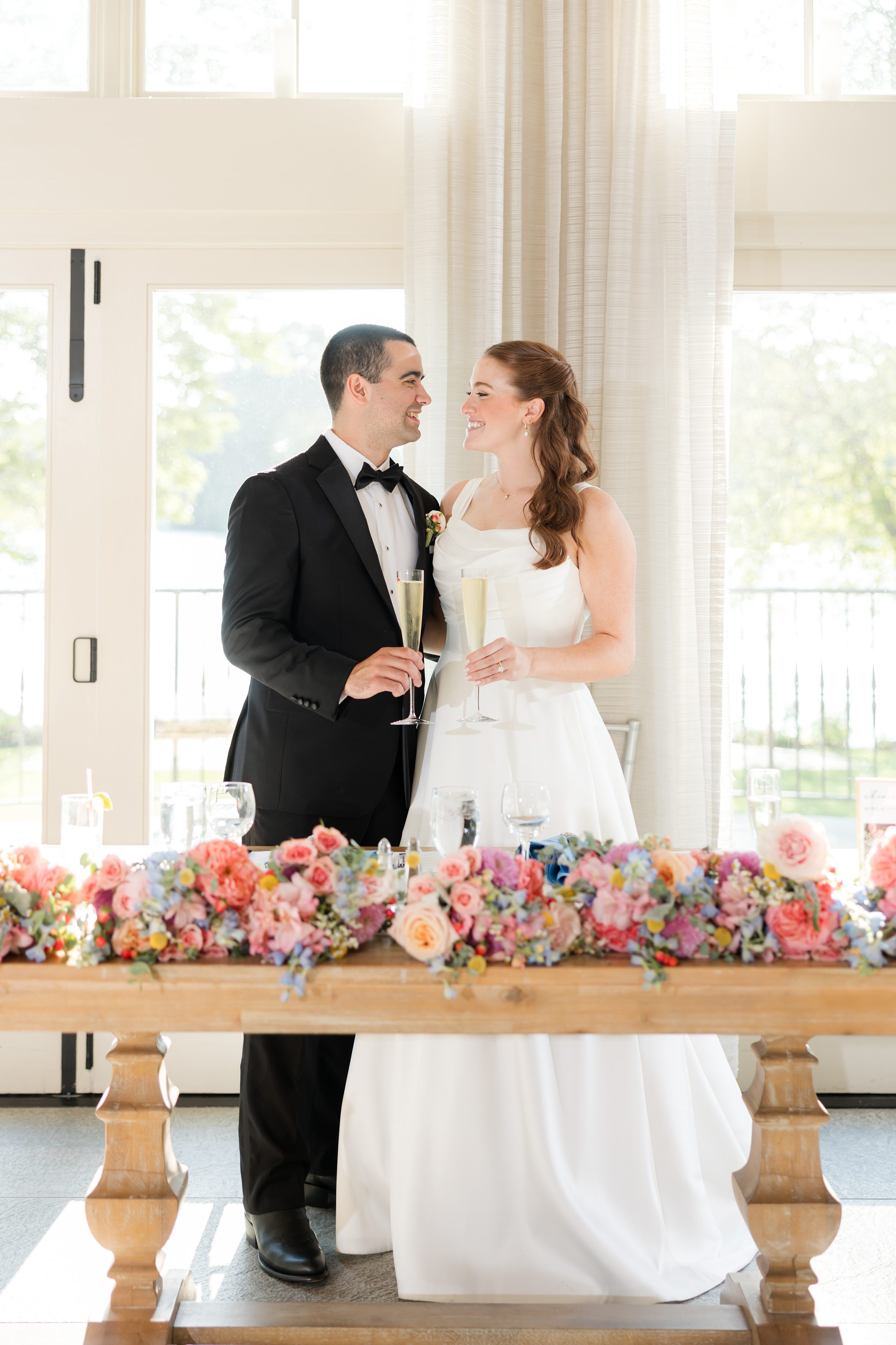 bride and groom looking at each other while holding a glass of champagne by the table filled with orange, powder blue, red flowers 
