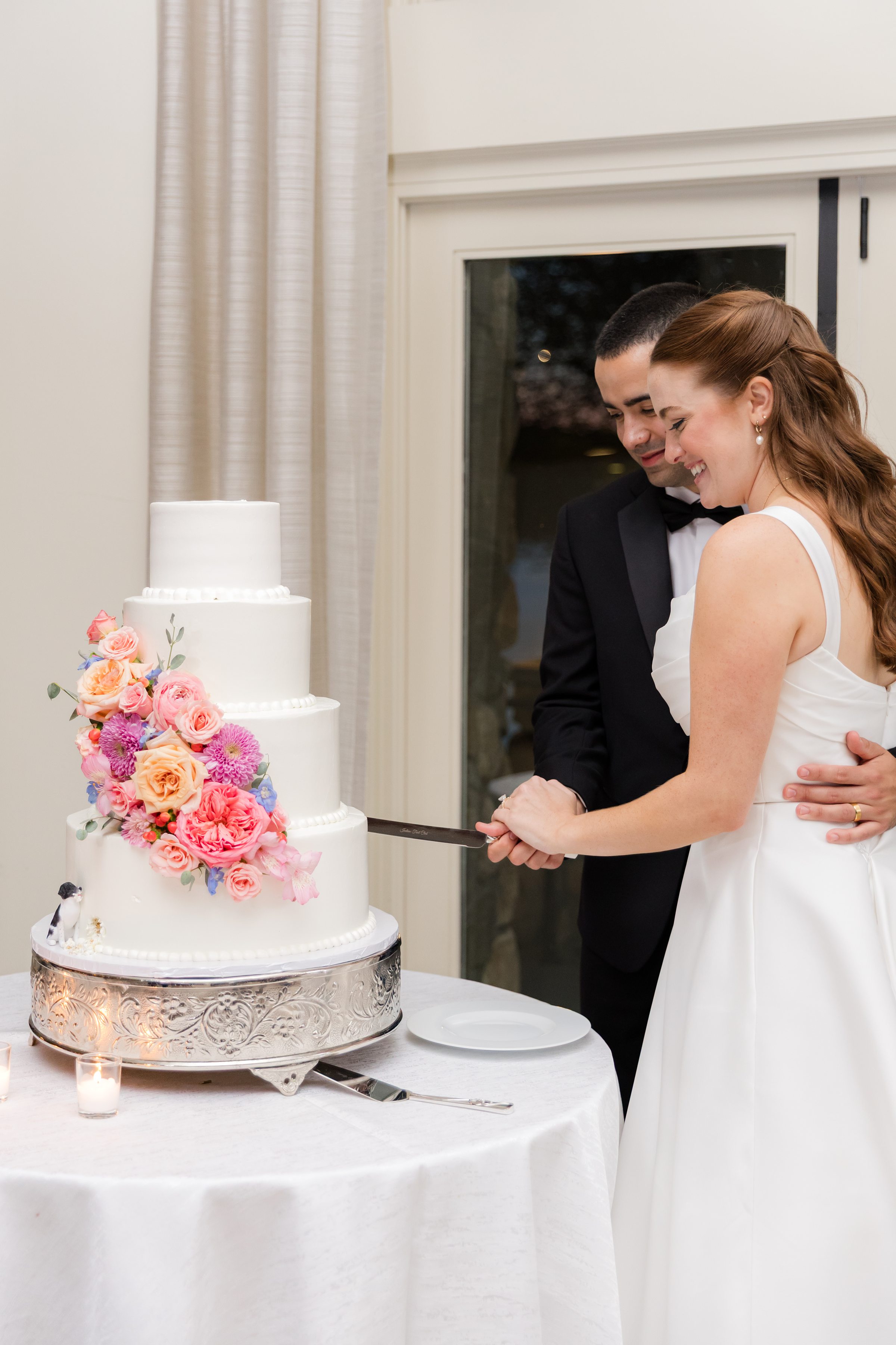 bride and groom slicing the white 4-tier cake with pink, orange, lavender, yellow flowers, together while smiling