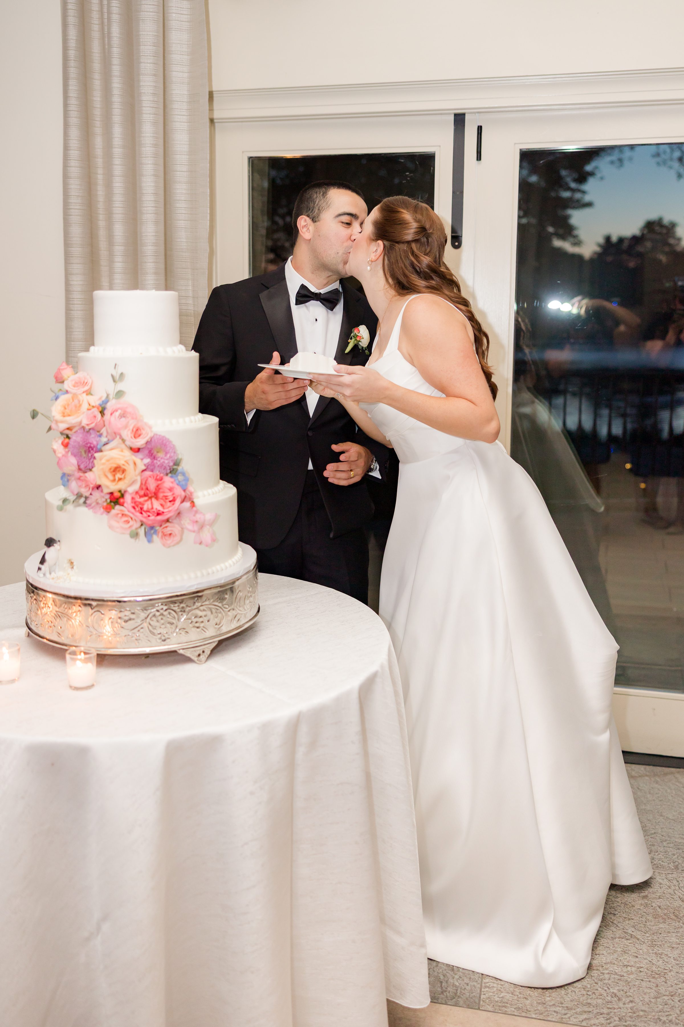 bride and groom kissing each other by the 4-tier wedding cake with pink, yellow, orange, lavender, powder blue glowers while holding a slice of cake on a plate