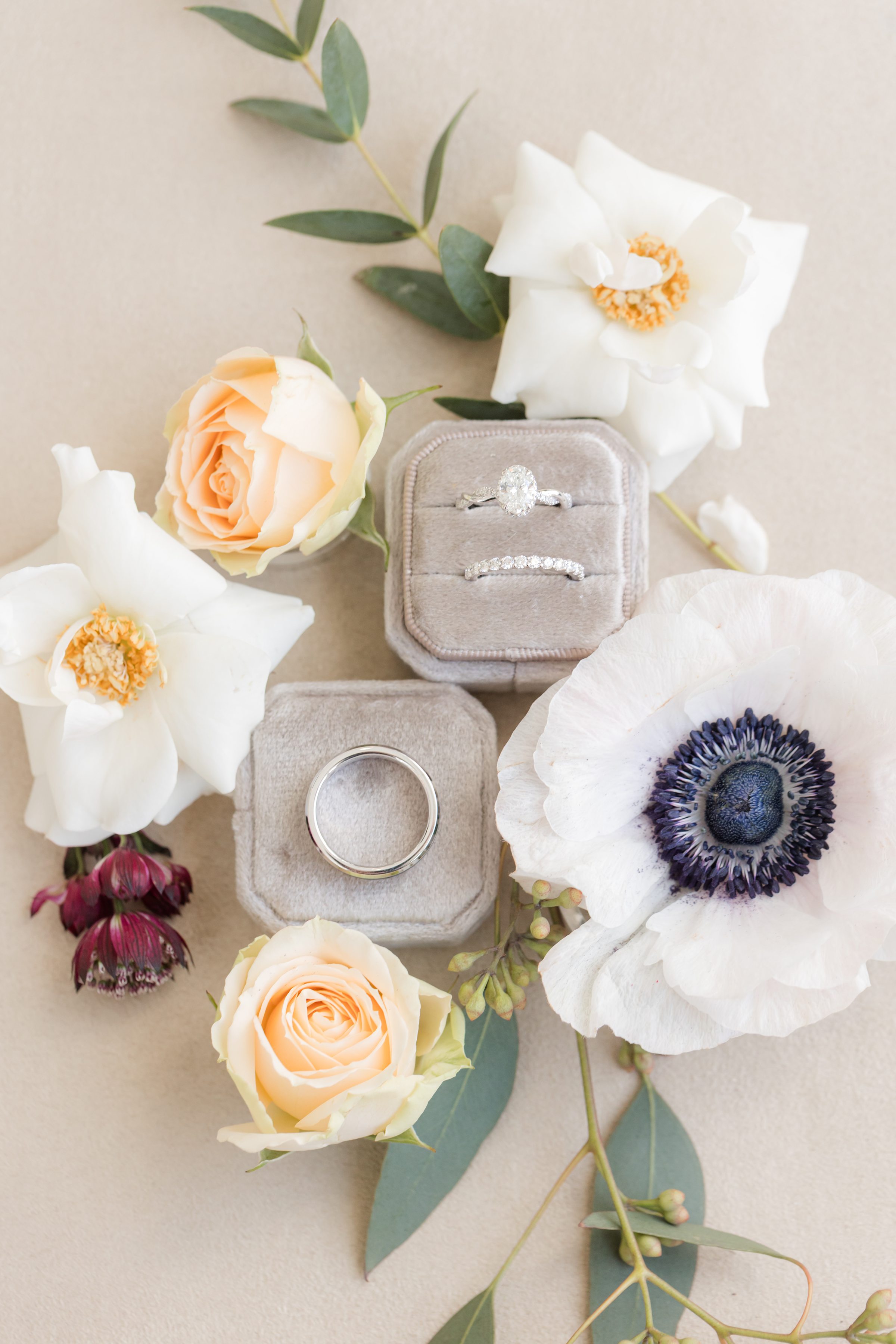Wedding rings in velvet boxes surrounded by white and blush flowers on a neutral background.