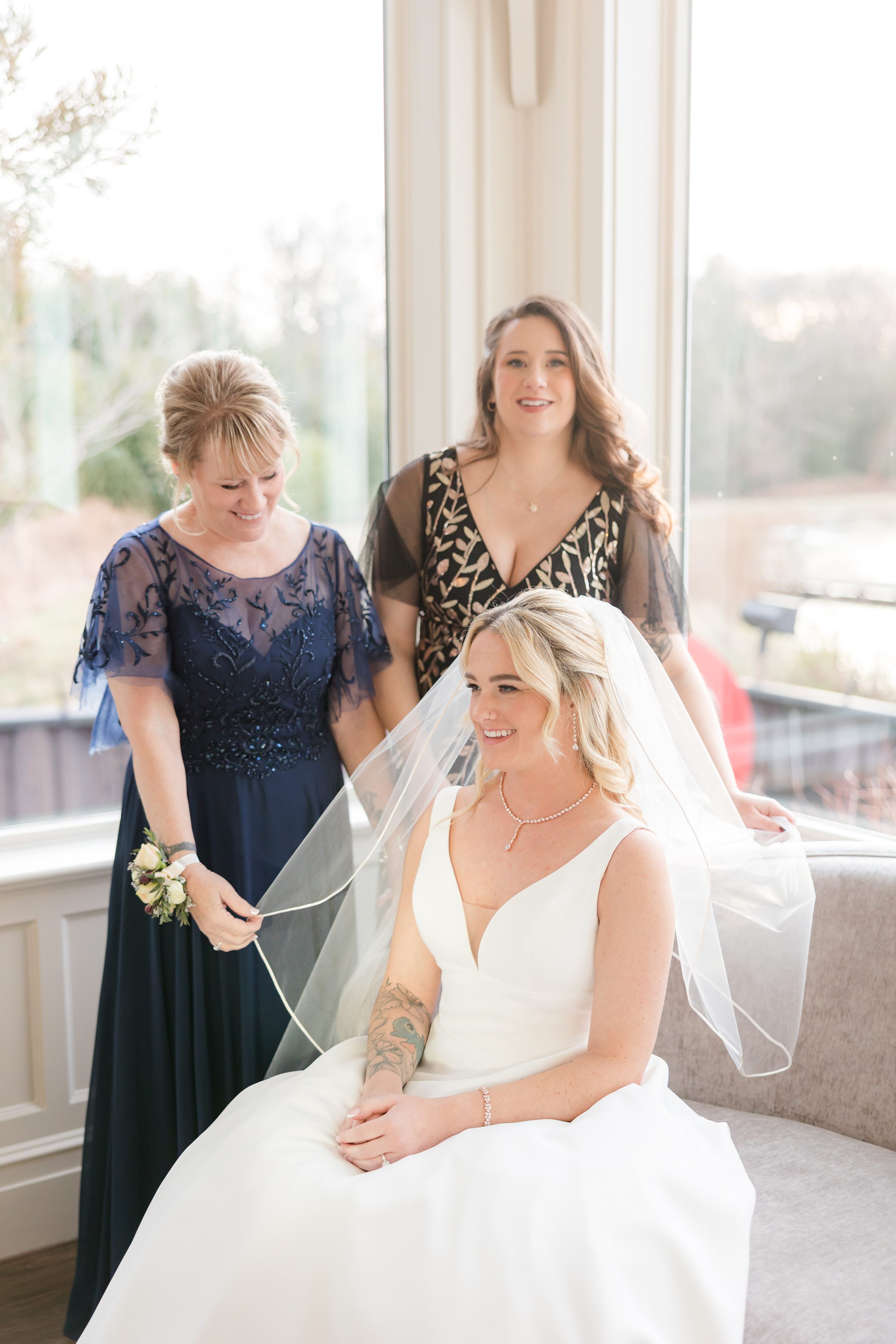 Bride sitting by a window smiling as her mother and sister adjust her veil in a bright, softly lit room.