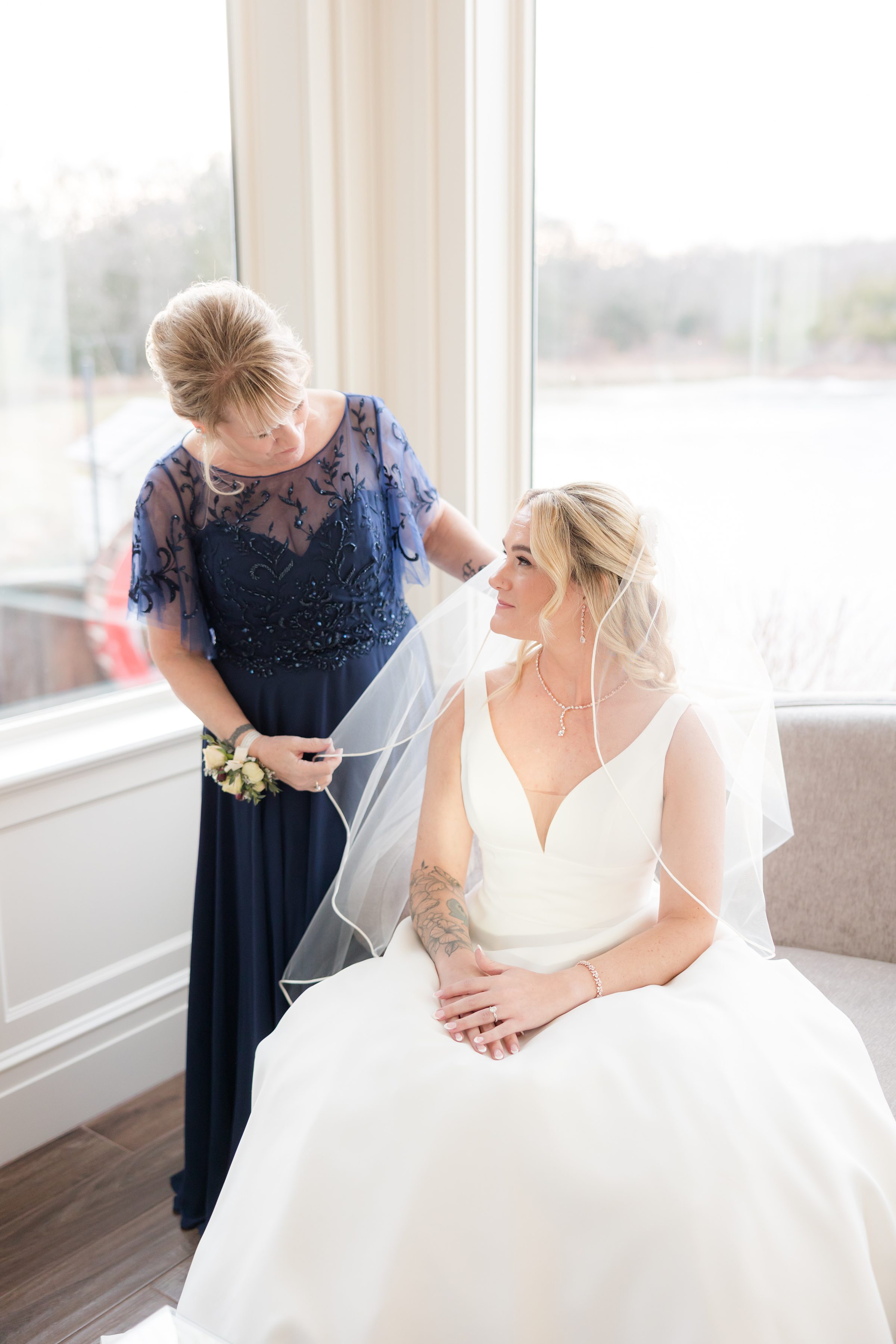 Bride seated by a window looking up as her mother gently adjusts her veil in soft natural light.