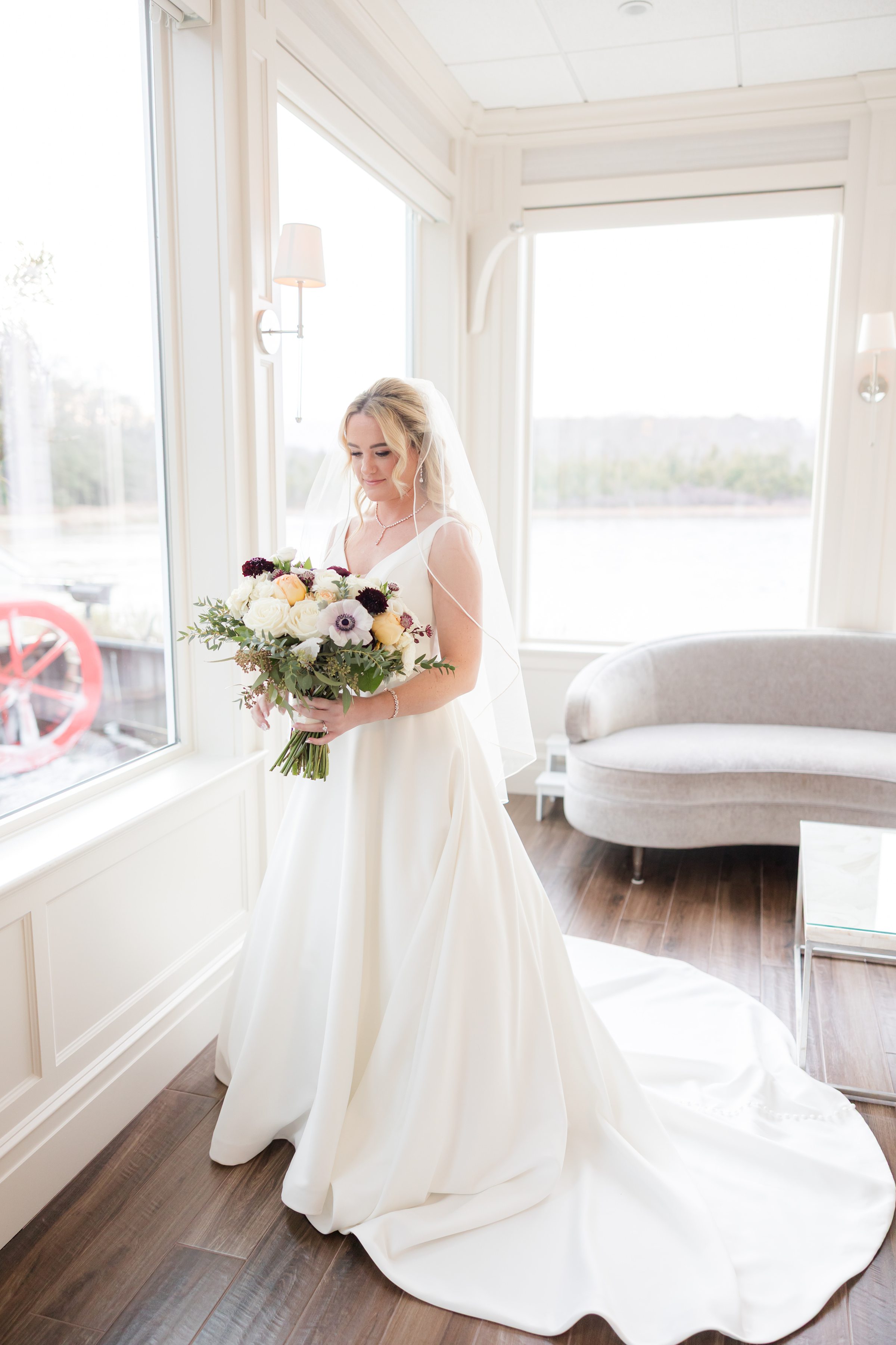 Bride in a white gown holding a bouquet by a large window with water view.