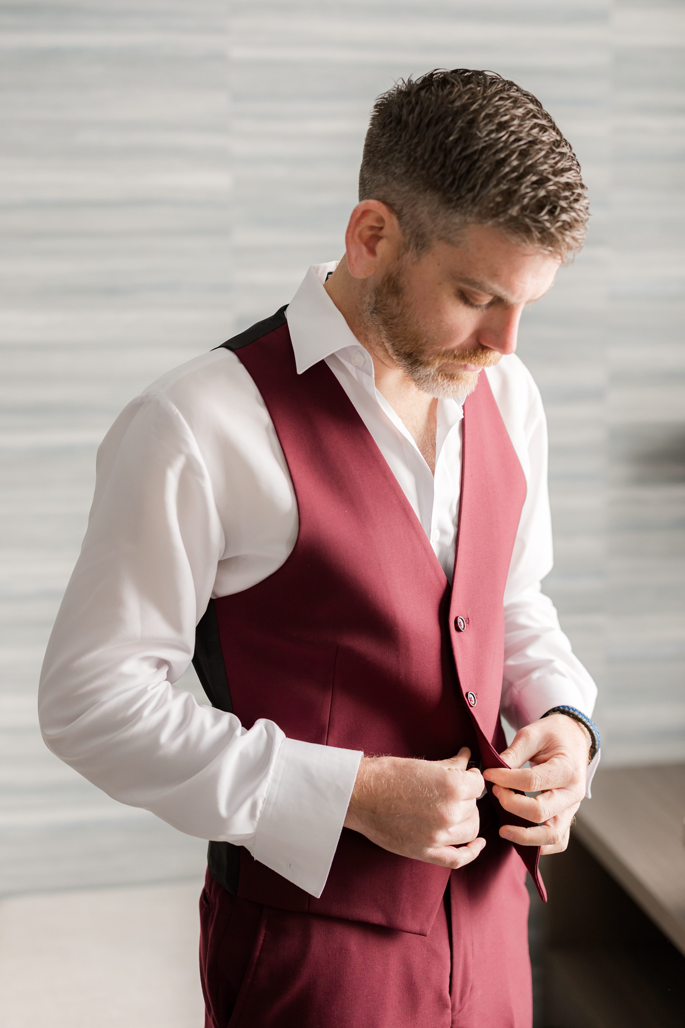 Groom in a burgundy vest looking down as he buttons his shirt in a softly lit room.
