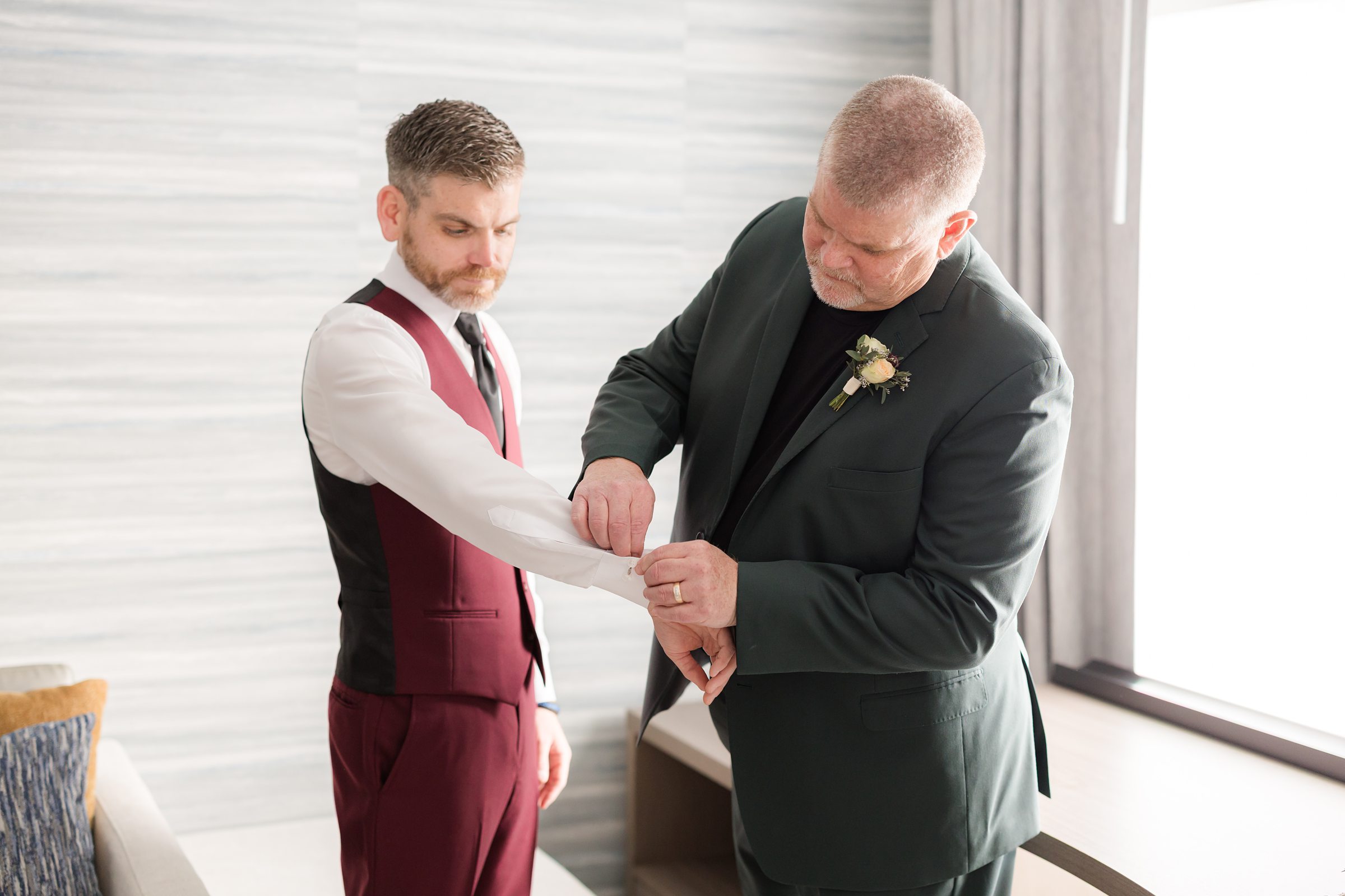 Groom standing with his father as his father adjusts his cufflinks in a hotel room.